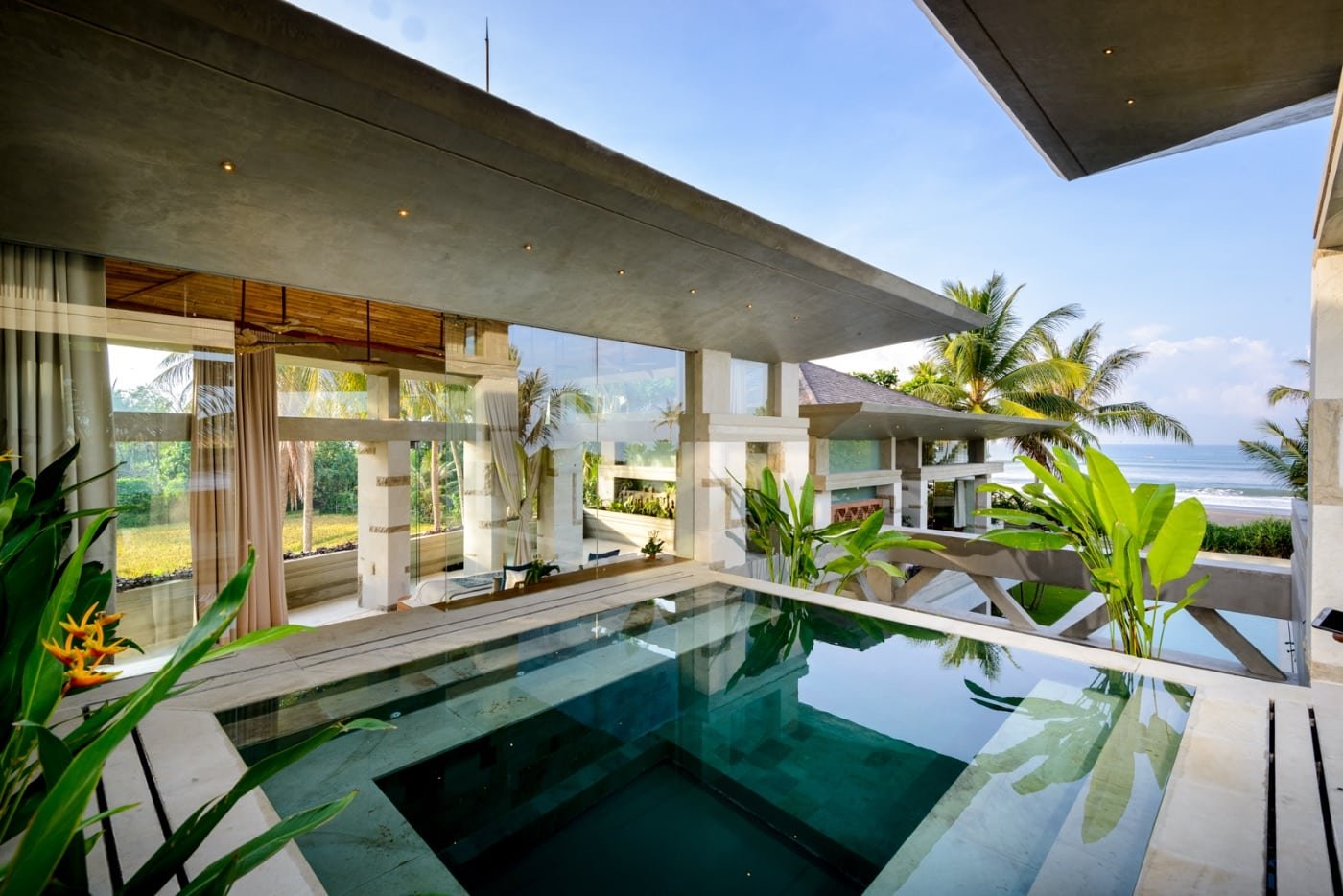 Modern indoor pool with large glass windows overlooking the beach, tropical plants, and palm trees outside.
