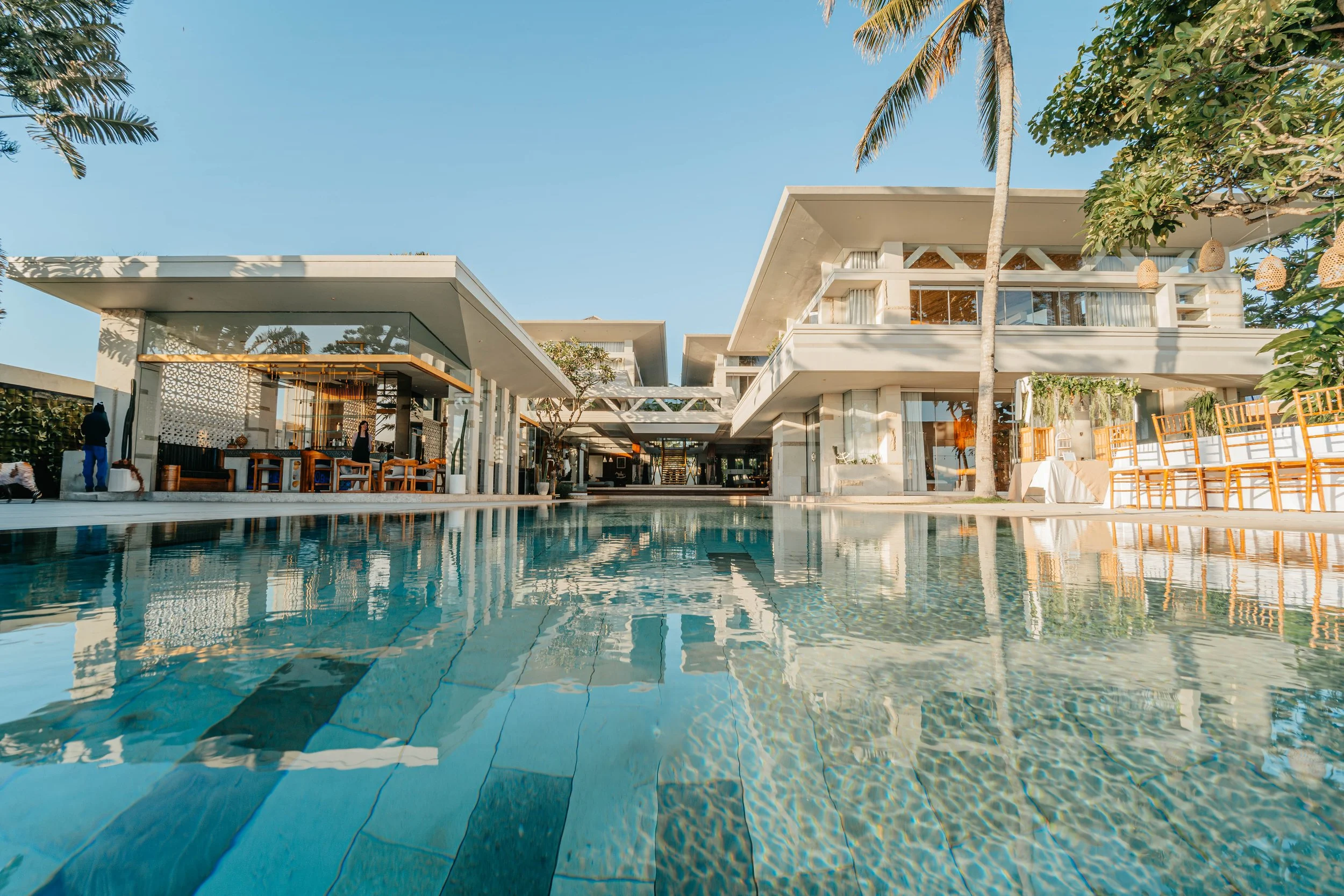Luxury resort with a large swimming pool, modern white buildings, palm trees, and outdoor seating areas under a clear blue sky.