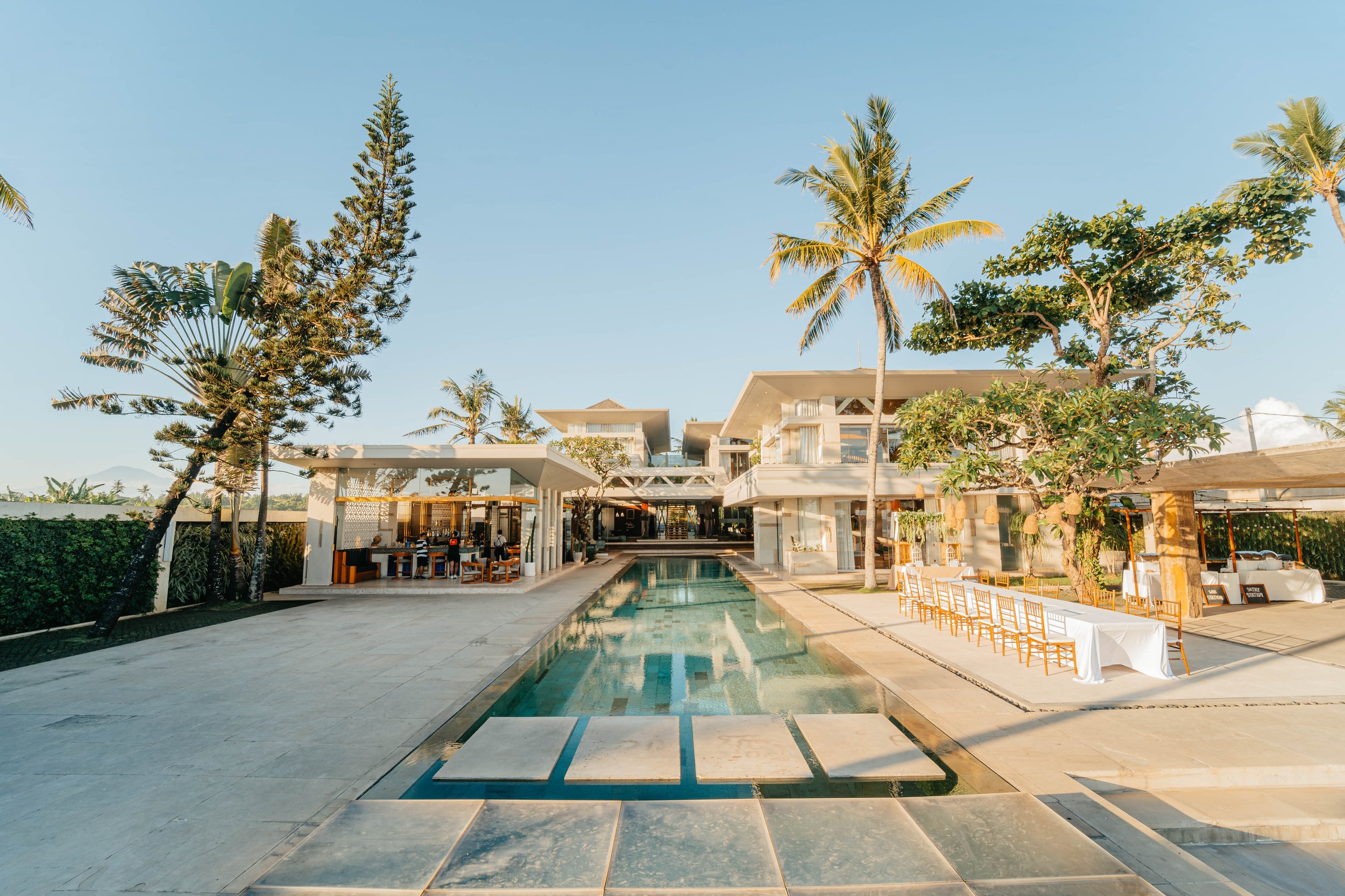 Luxury resort with a modern building, swimming pool, and tropical trees under a clear sky.