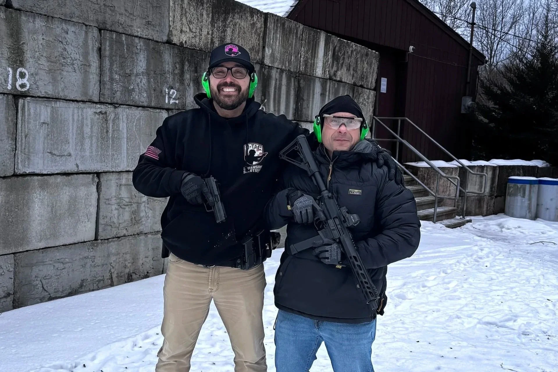 Two men in winter clothing and ear protection holding guns outdoors in the snow, standing in front of a stone wall and a building.