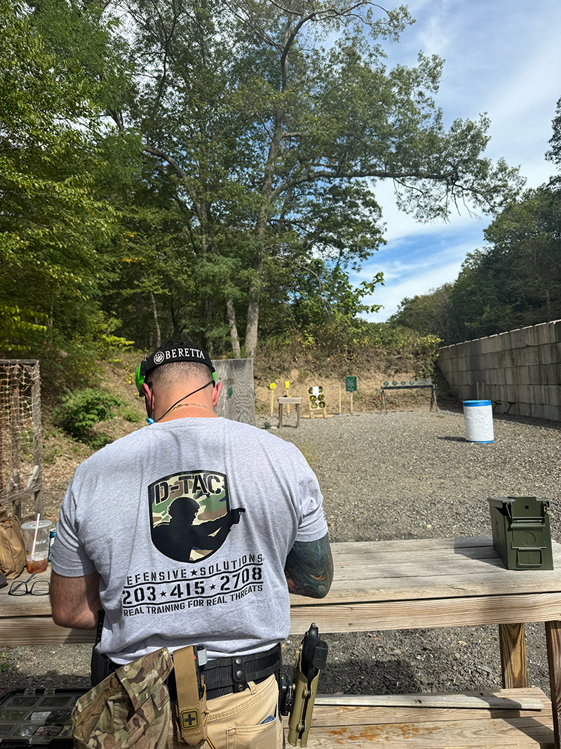 Man at outdoor shooting range with target stands, barrels, and trees in background, wearing a gray shirt, black hat, and tactical gear.