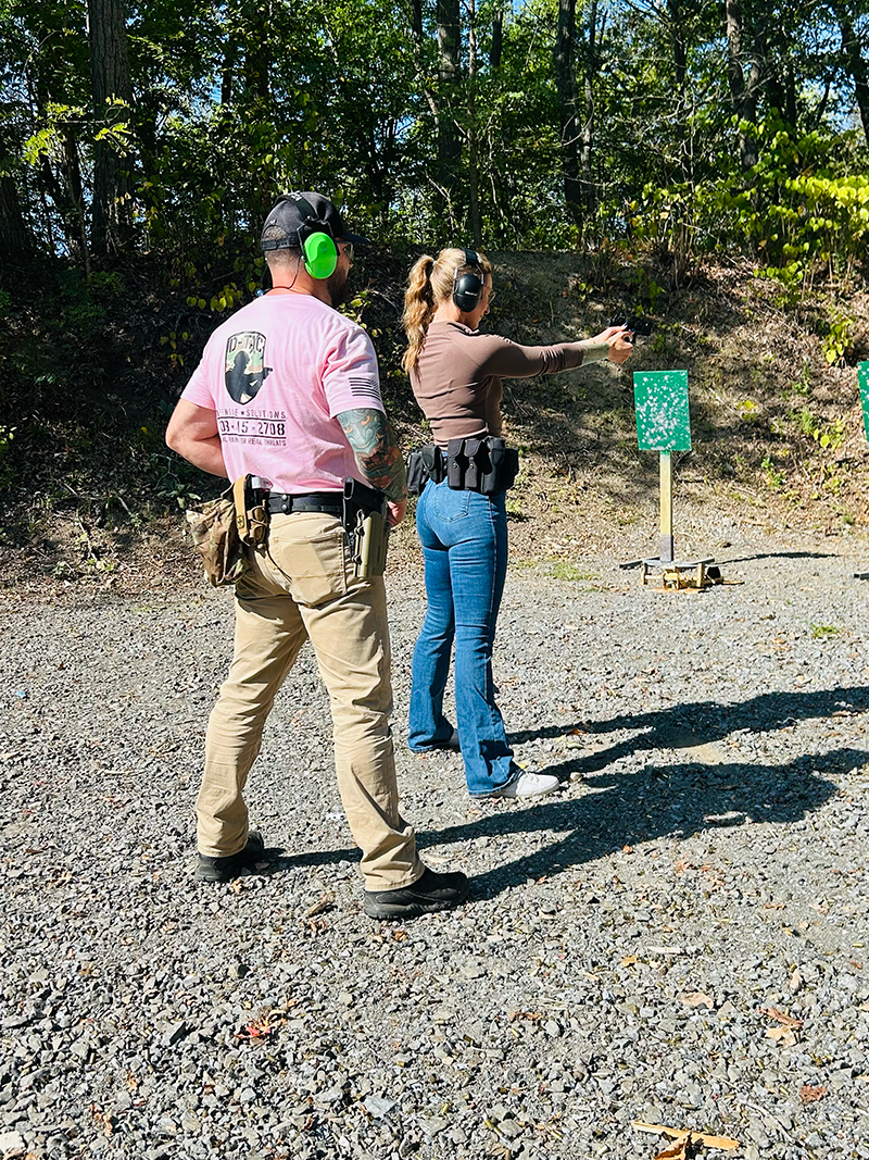 Two people at an outdoor shooting range with trees in the background, wearing hearing protection and standing on gravel with a green target in front of them.