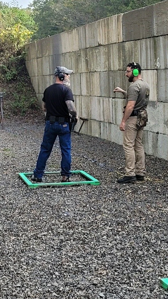 Two men at an outdoor shooting range, one inside a green rectangular frame on the ground, the other beside him, both wearing hearing protection and casual tactical clothing, next to a concrete wall.