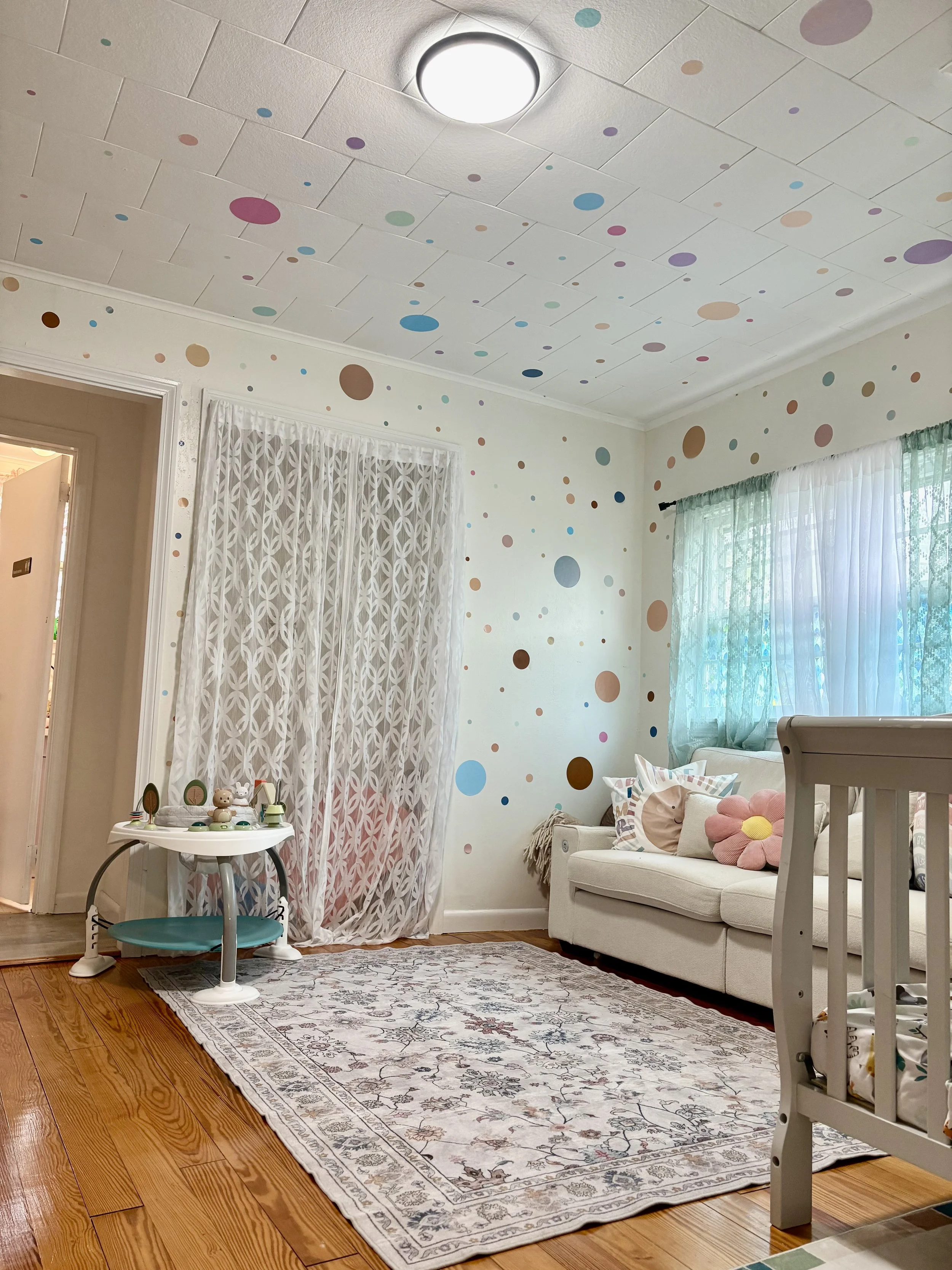 A cozy living room with wooden flooring, a vintage-looking rug, a white sofa with decorative pillows, sheer curtains over windows, a small table with plush toys, and colorful polka dot wall and ceiling decorations.