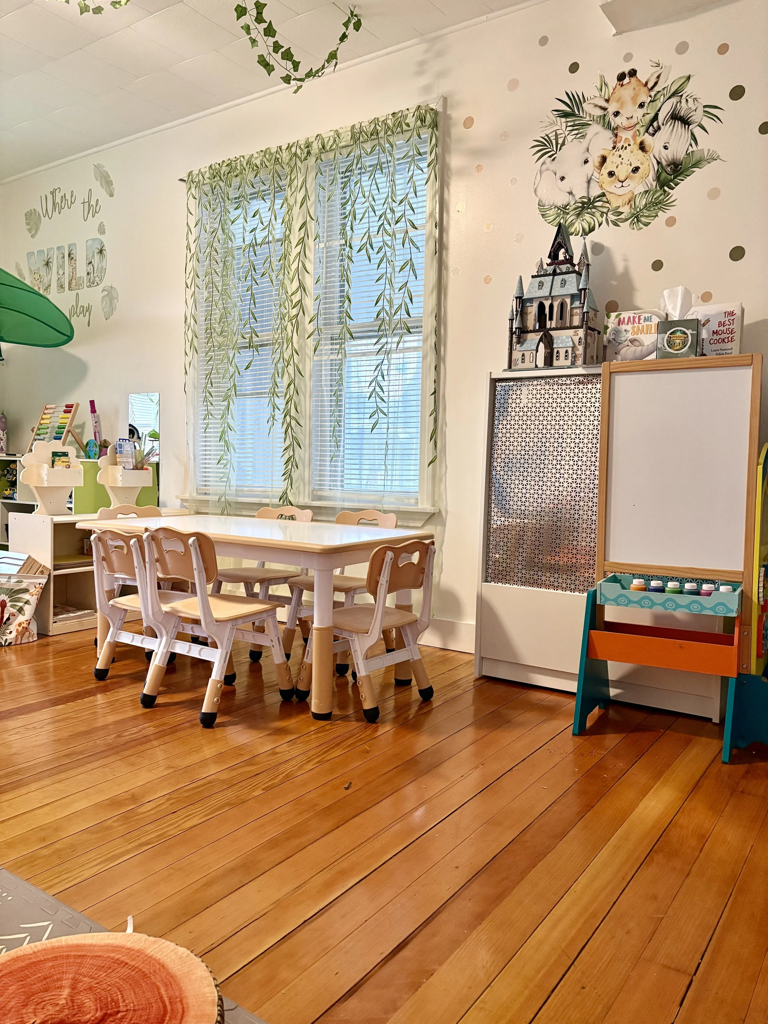 A corner of a children's playroom with a wooden floor, a small table surrounded by chairs, a bookshelf, and various toys and decorations including wall art with animals and a castle model on top of a white cabinet.