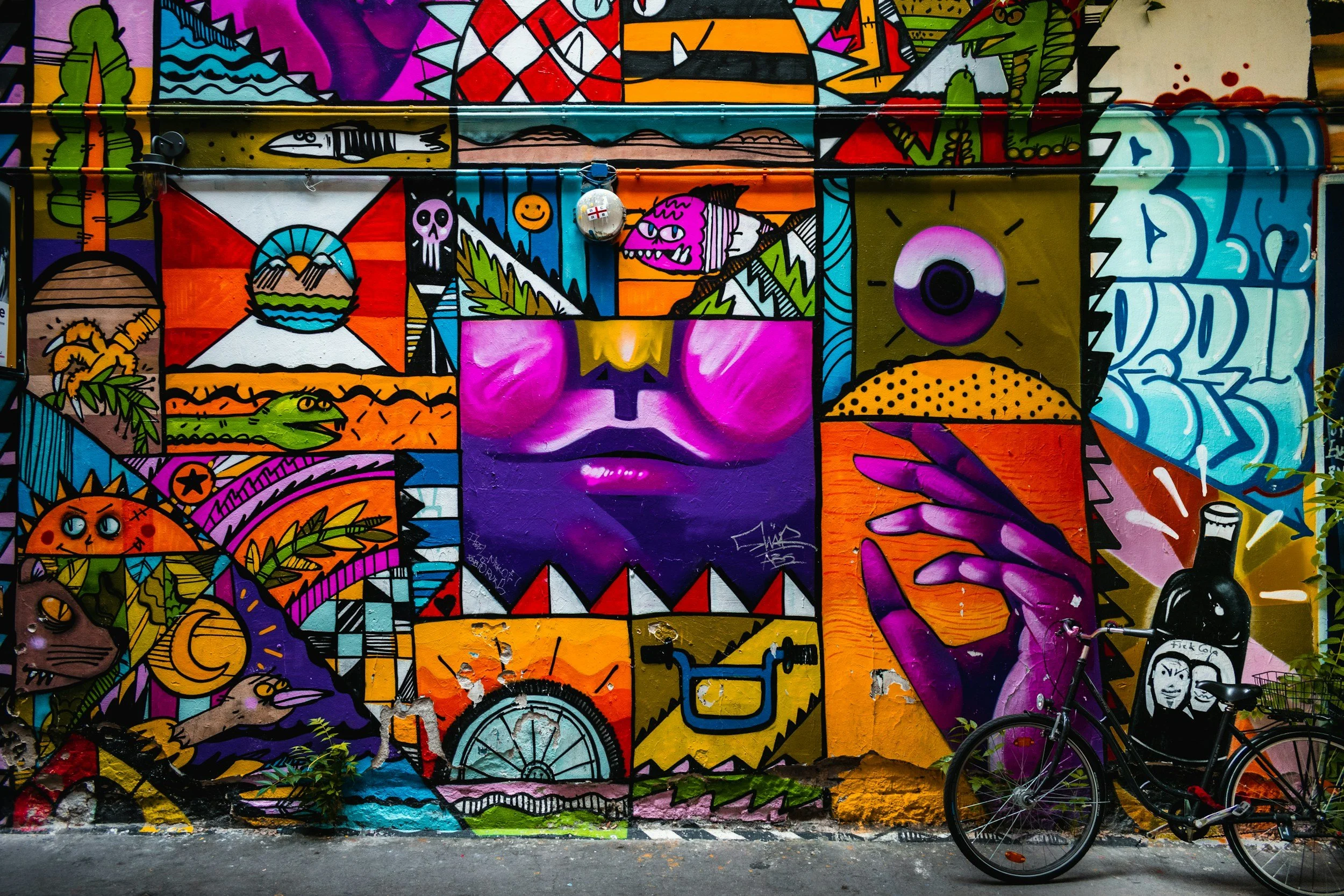 Colorful mural painting featuring various abstract and surreal elements including a large purple face with closed eyes in the center, a hand reaching from the right side, an eye in the upper right corner, a fish, a skull, a mountain landscape, and various geometric shapes and characters, with a bicycle parked in front.