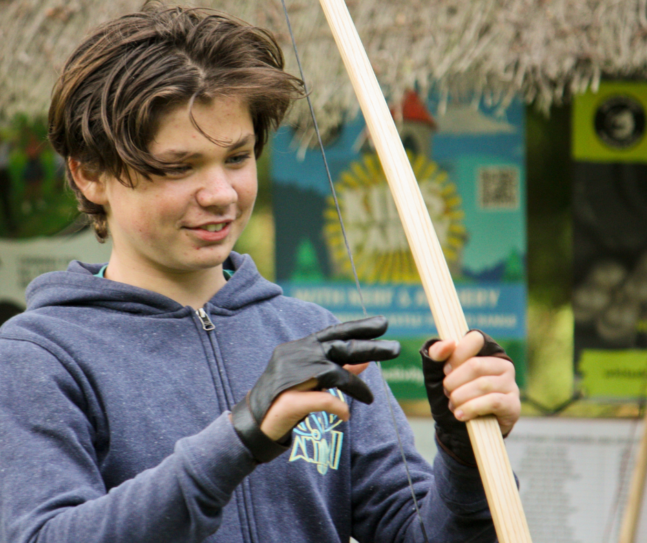 A young boy with brown hair smiling, wearing a blue hoodie and black gloves, holding a wooden stick with a string attached, outdoors with colorful banners in the background.