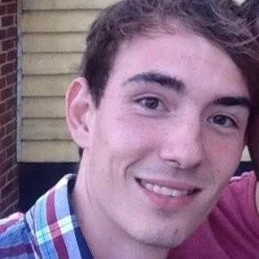 Close-up of a young man smiling outdoors, wearing a colorful plaid shirt.