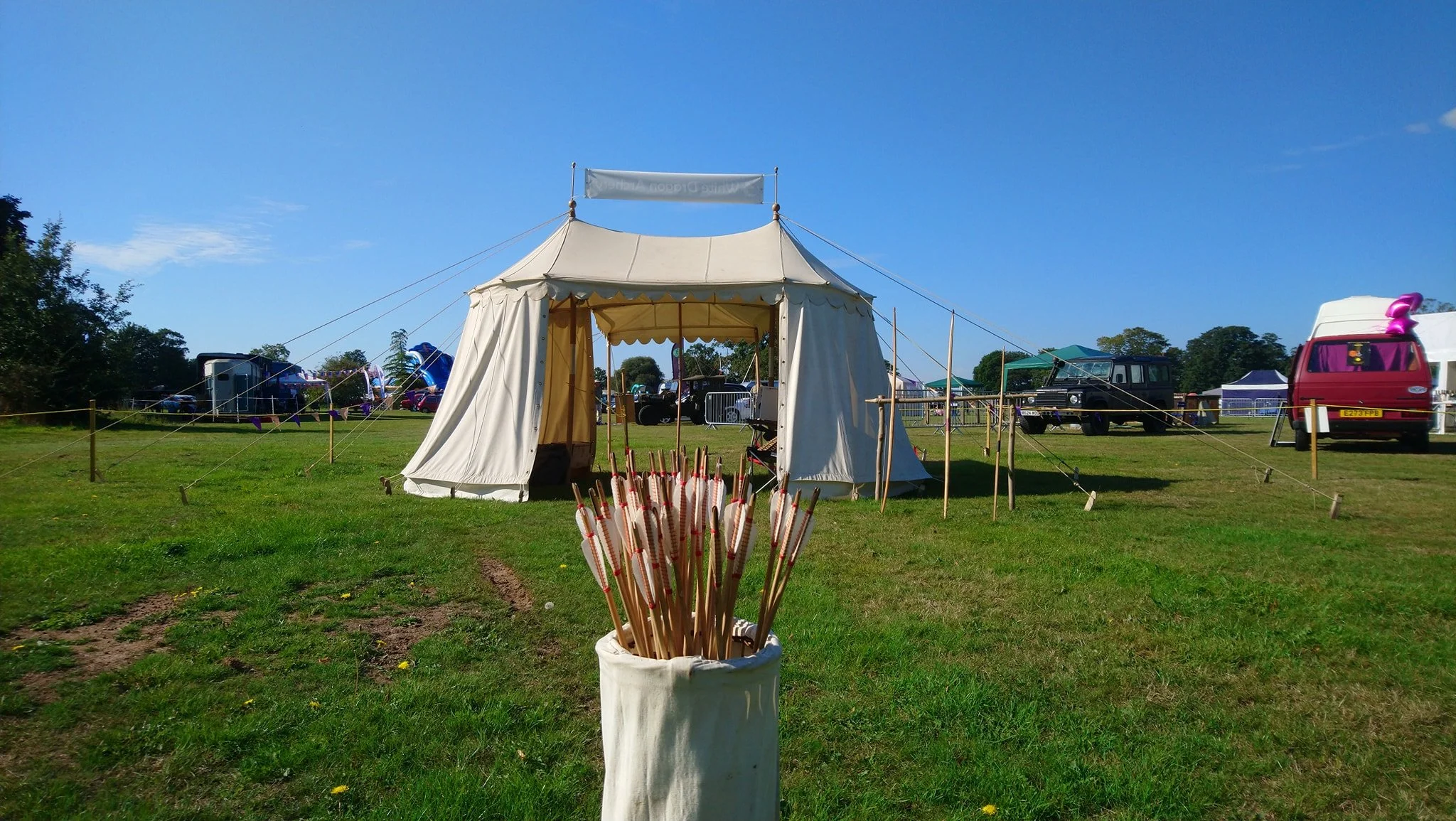A white event tent set up in a grassy field with a cluster of arrows in a white bag in the foreground, and several vintage vehicles and tents in the background.