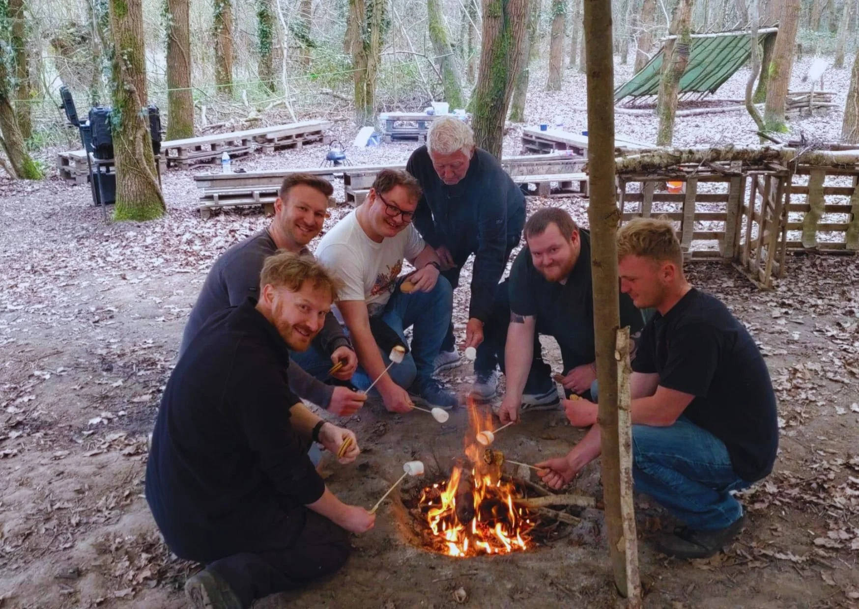 Group of seven men gathered around a campfire roasting marshmallows in a wooded outdoor area.