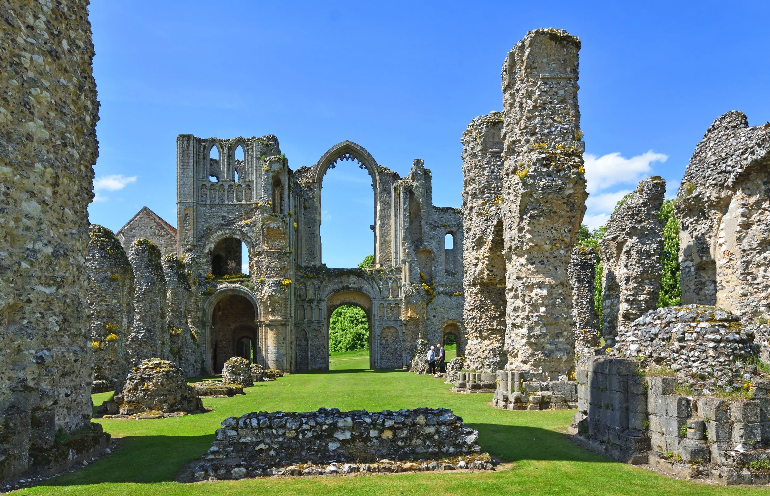 Bury St Edmunds Abbey Ruins is an ancient stone building with tall, weathered walls and arches, surrounded by a grassy area and a blue sky with some clouds.