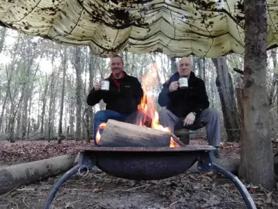 Two men toasting the camera with cups of tea as they sit by a campfire in a wood, beneath a khaki army parachute