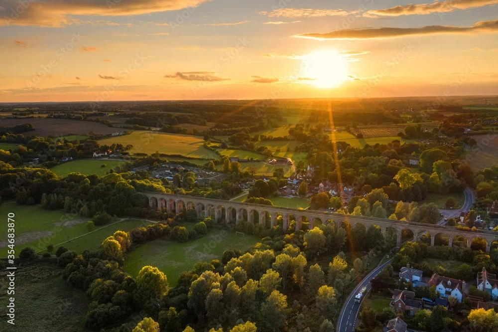 An aerial view of Chelmsford at sunset, featuring a large stone viaduct, green fields, trees, and a few scattered houses.