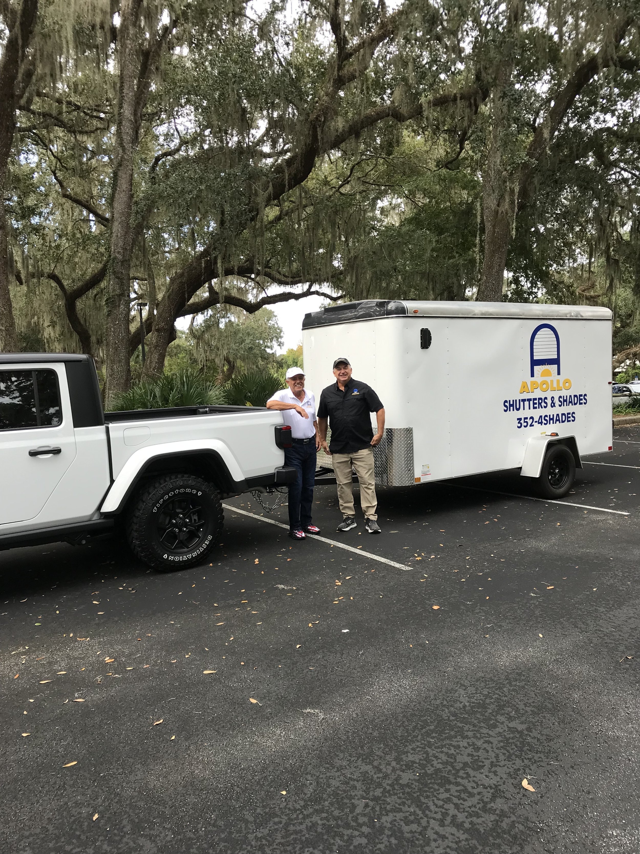 Two men standing next to a white truck and a trailer with 'APOLLO SHUTTERS & SHADES' written on it, parked in a lot with large trees in the background.