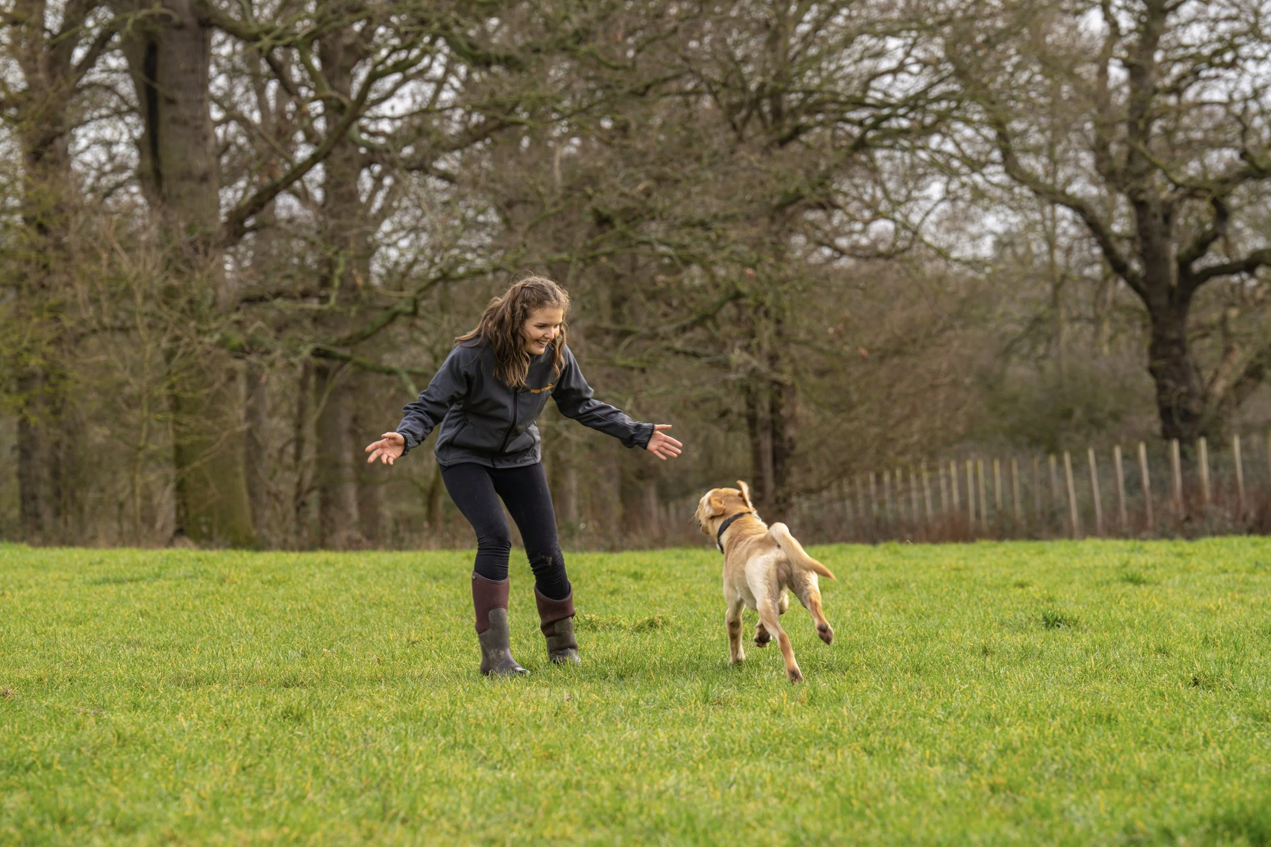 A woman playing fetch with a dog in a grassy field with trees in the background.