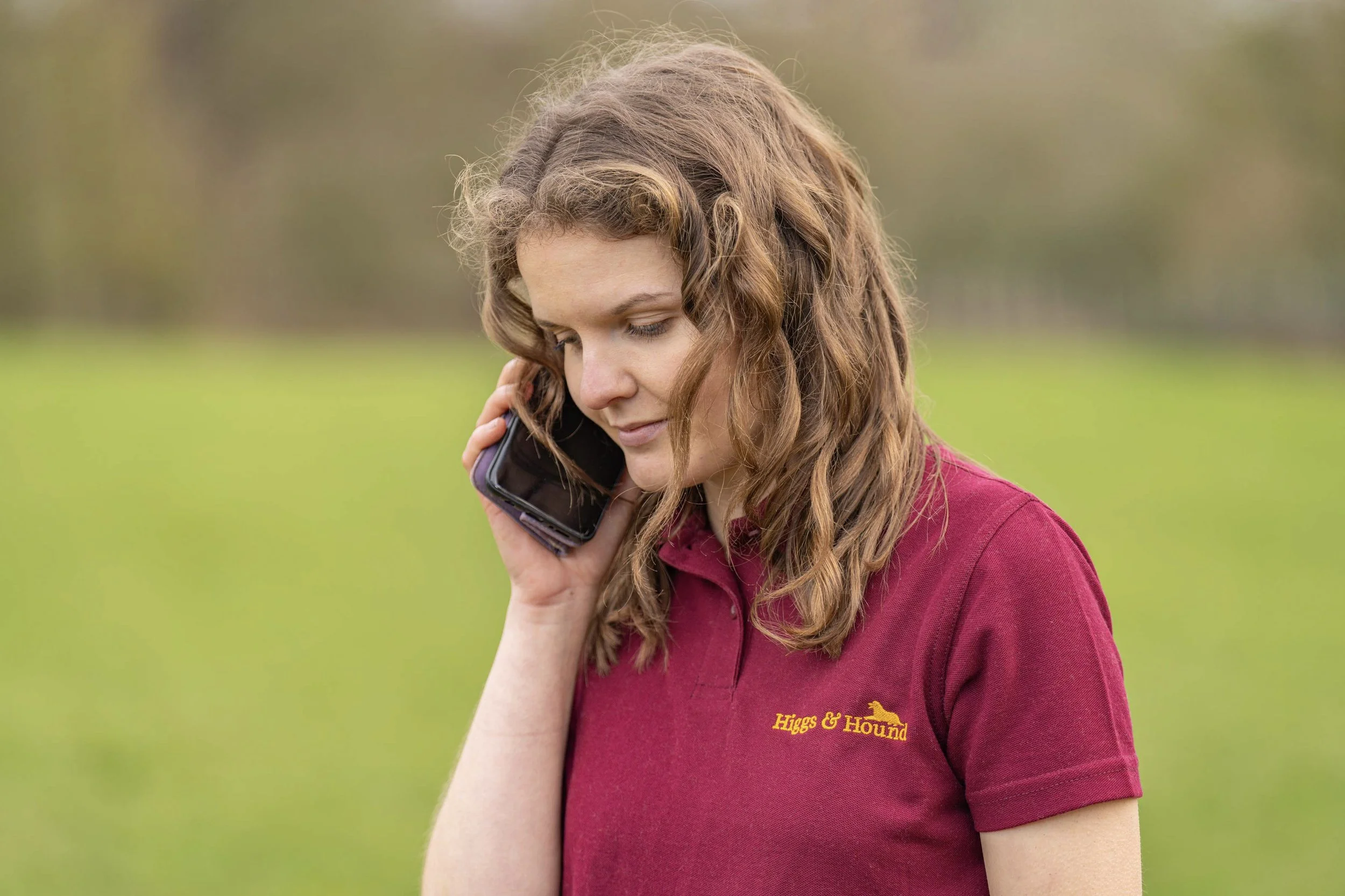 Alice with curly brown hair wearing a maroon collared shirt talking on a black mobile phone outdoors on a grassy field.