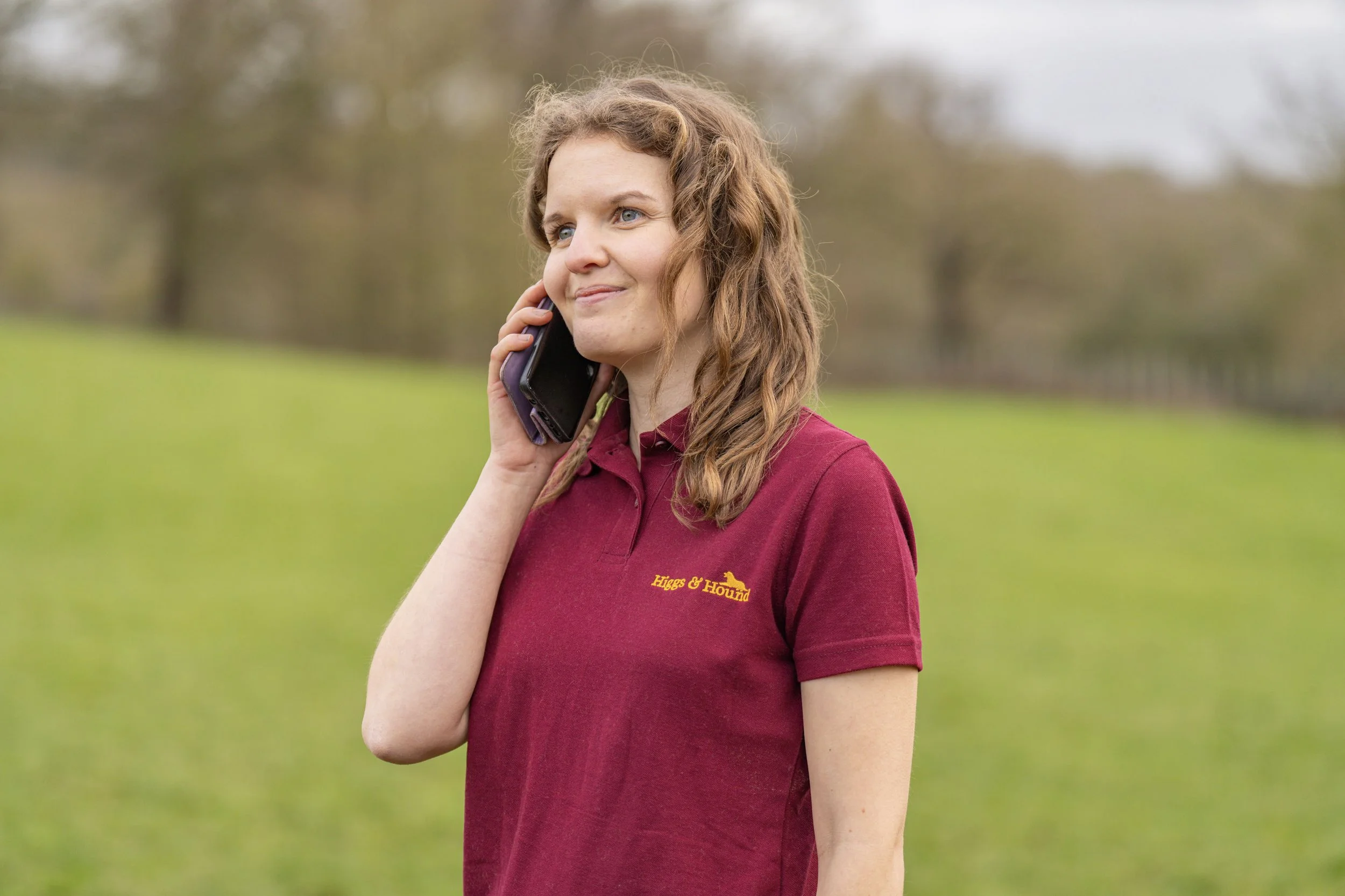 A young woman with curly, reddish-brown hair talking on her cell phone outdoors in a grassy field with trees in the background, wearing a maroon polo shirt with a yellow logo that says "Higgs & Hound."