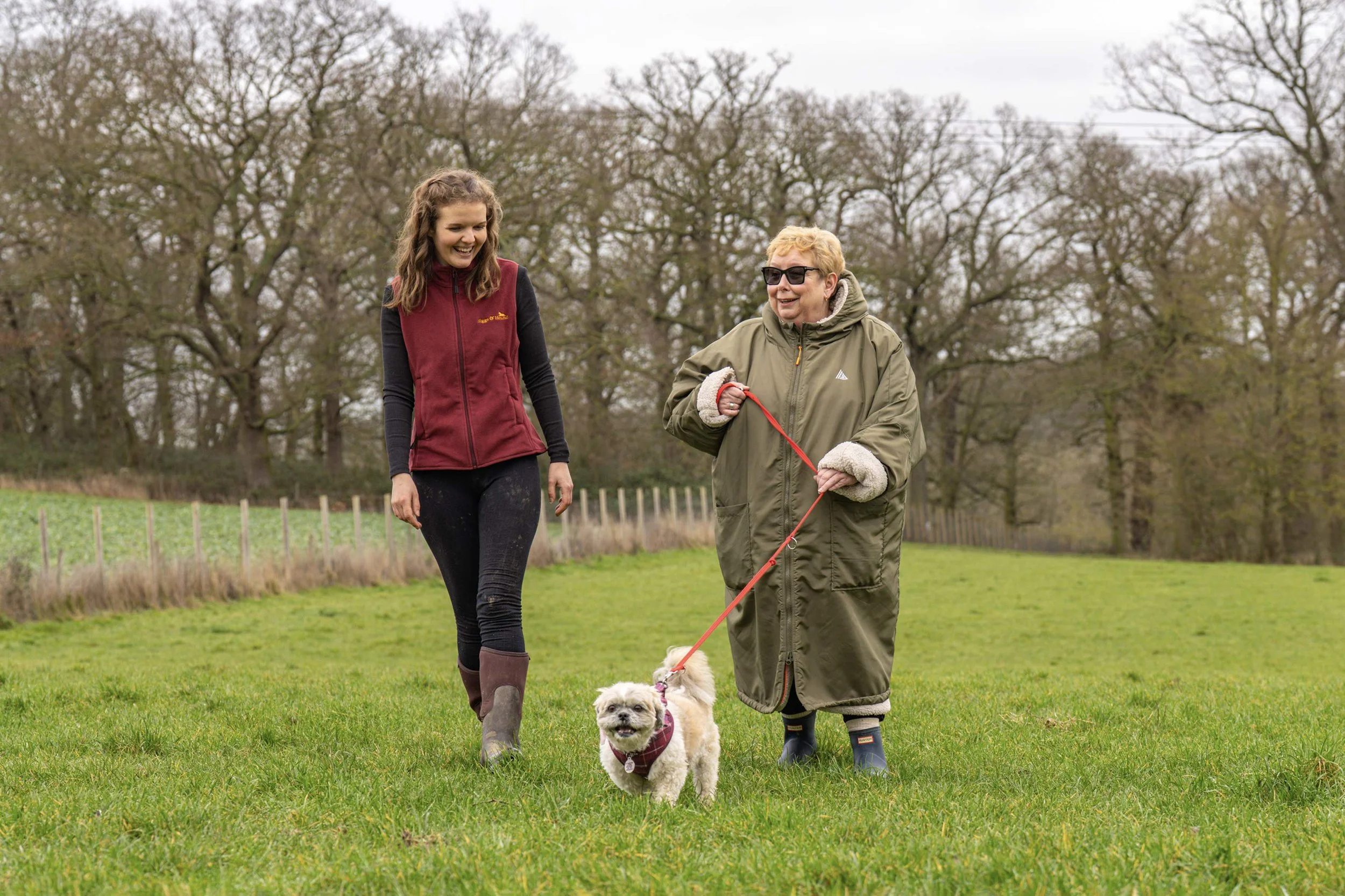 Two women (Alice and her client) walking and talking in a grassy park with their dog.