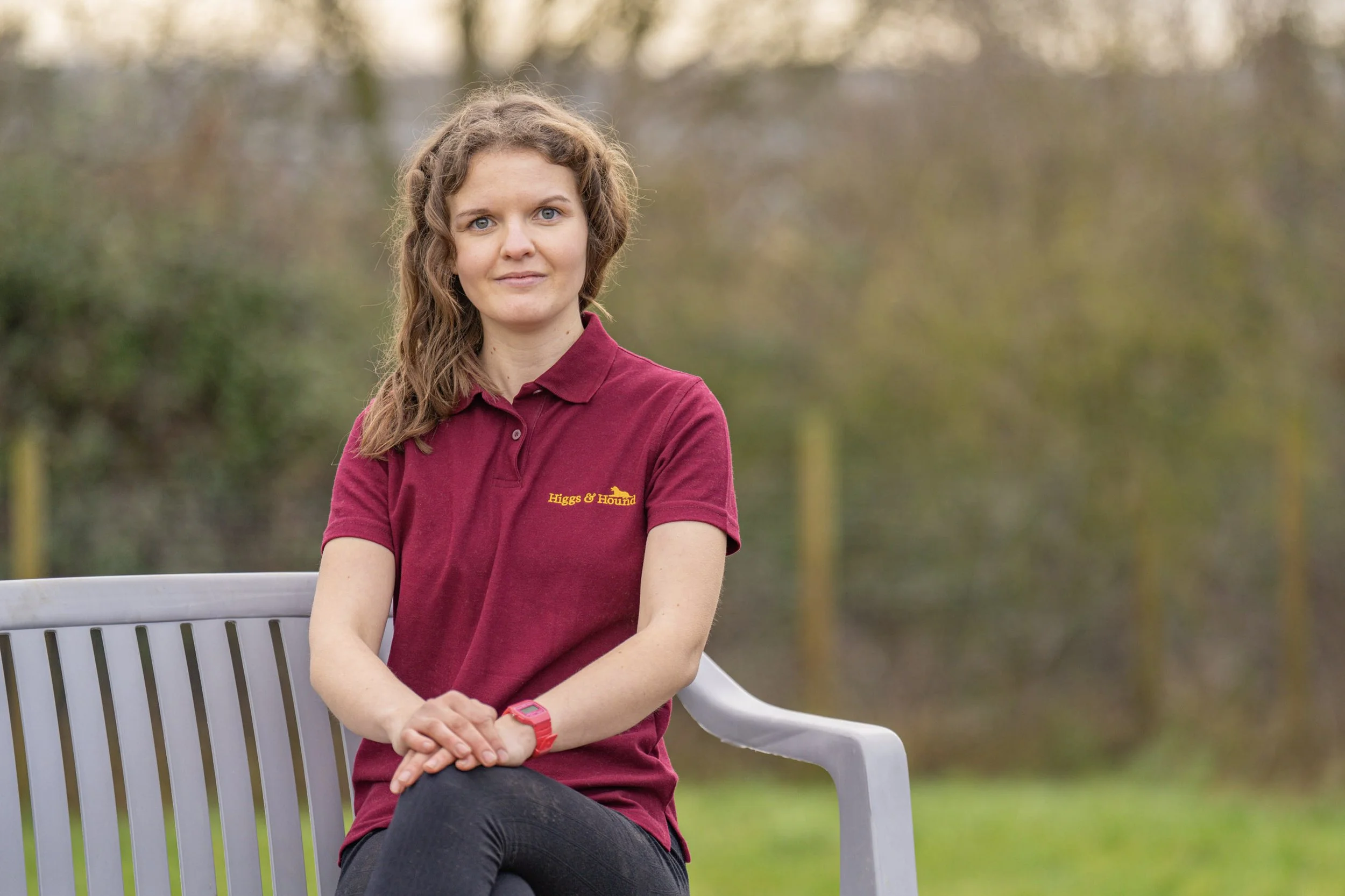 A woman with wavy brown hair, wearing a maroon polo shirt with yellow embroidery, sitting on a white park bench outdoors, looking at the camera with a neutral expression.
