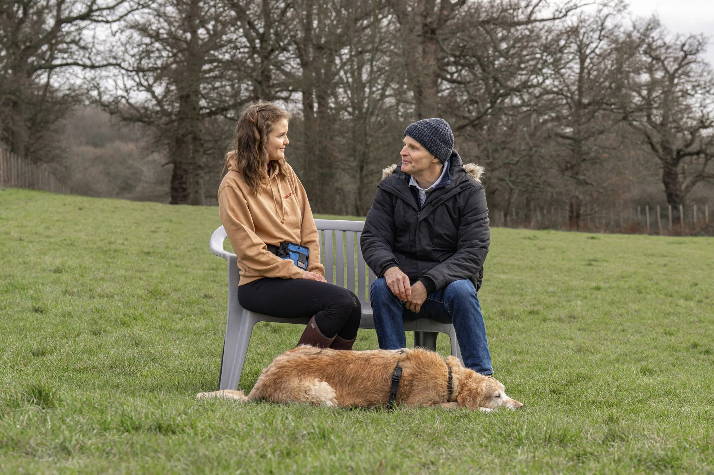 A young woman and a man sit on a gray bench outdoors, talking to each other. A golden retriever lies on the grass in front of them. The background shows leafless trees and an overcast sky.