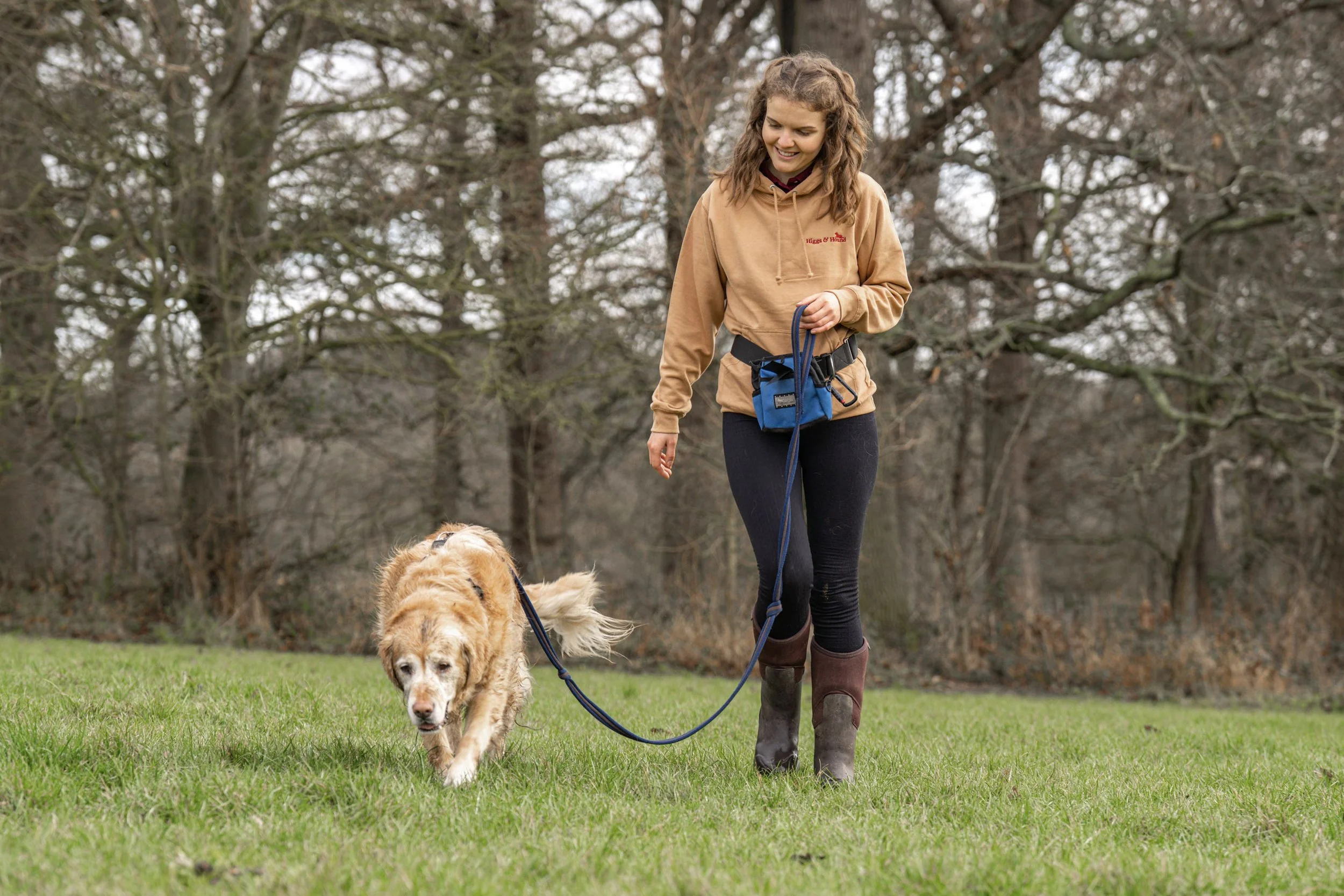 A woman walking a golden retriever on a leash in a grassy park with leafless trees in the background.