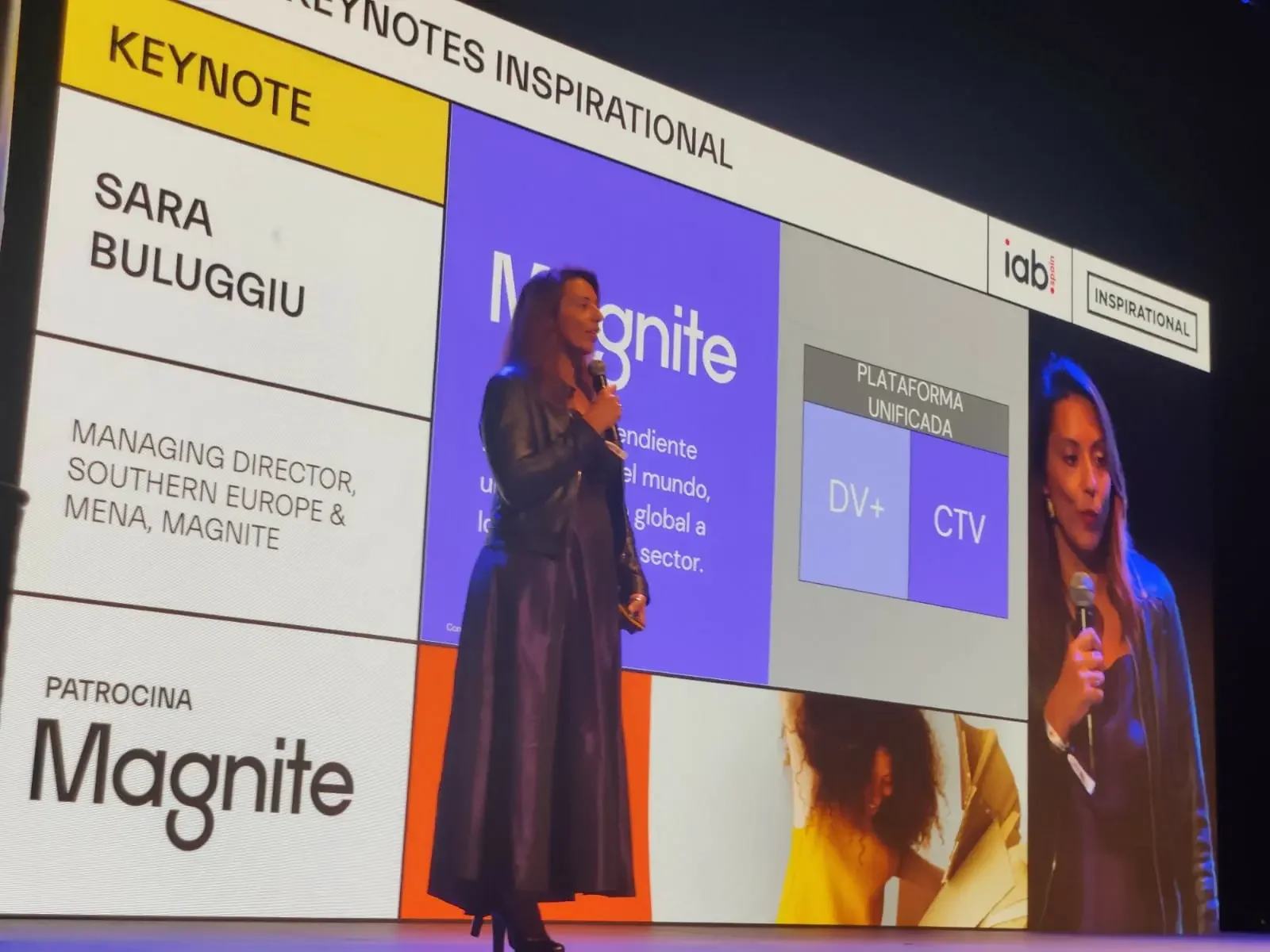 Donna con giacca nera che parla al microfono durante una presentazione, con uno schermo grande dietro di lei che mostra testo e grafici in italiano.