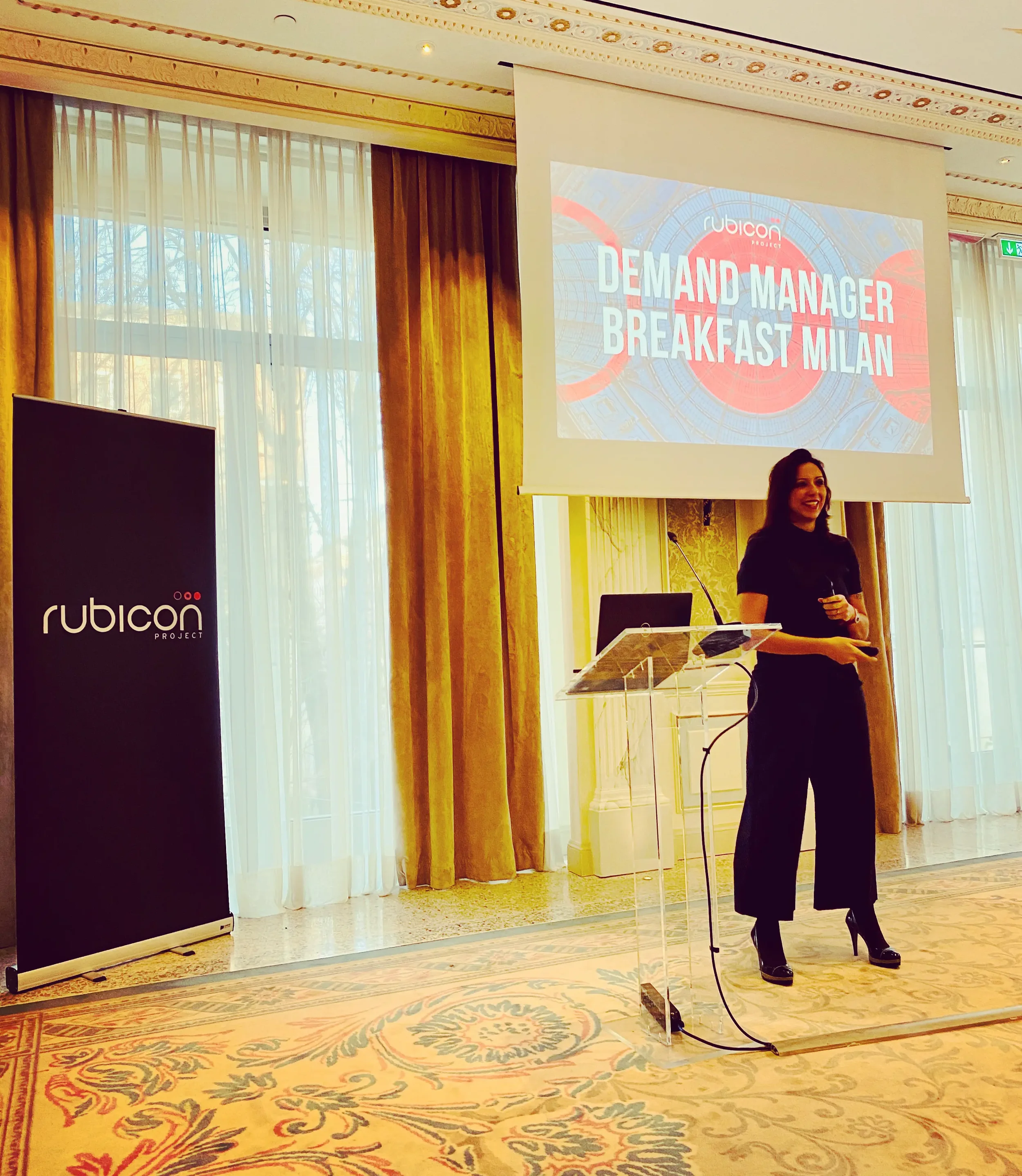 Una donna in piega elegante sta dando una presentazione in una sala decorata con tendaggi dorati. Sullo schermo dietro di lei si legge 'Demand Manager Breakfast Milan' con il logo 'Rubicon Project'. C'è anche un banner con lo stesso logo sulla sinistra.