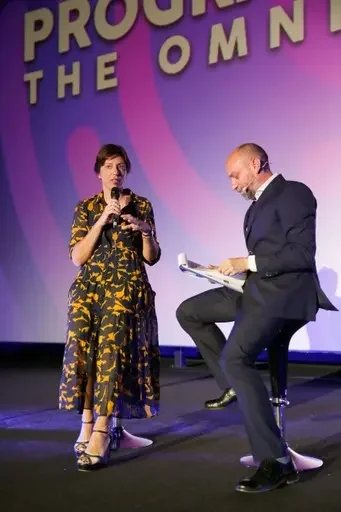 Due persone sul palco, una donna con un abito floreale e un uomo in giacca scura, che interagiscono durante un evento.