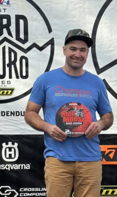 Smiling man wearing a black cap, blue Champion t-shirt, and khaki pants, holding a red and black award plaque with the text 'MS Muddy'. He stands in front of a backdrop with logos for Dirt Euro and other brands.