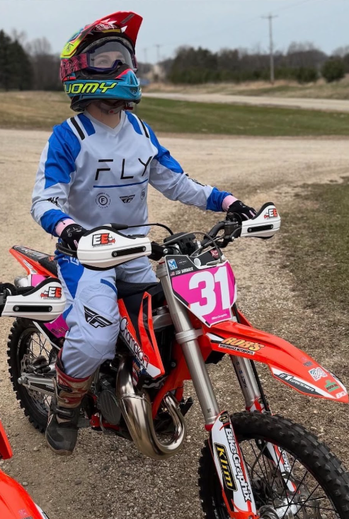 Child wearing a colorful helmet, goggles, and motocross gear sitting on a red dirt bike with the number 31, outdoors on a dirt track.