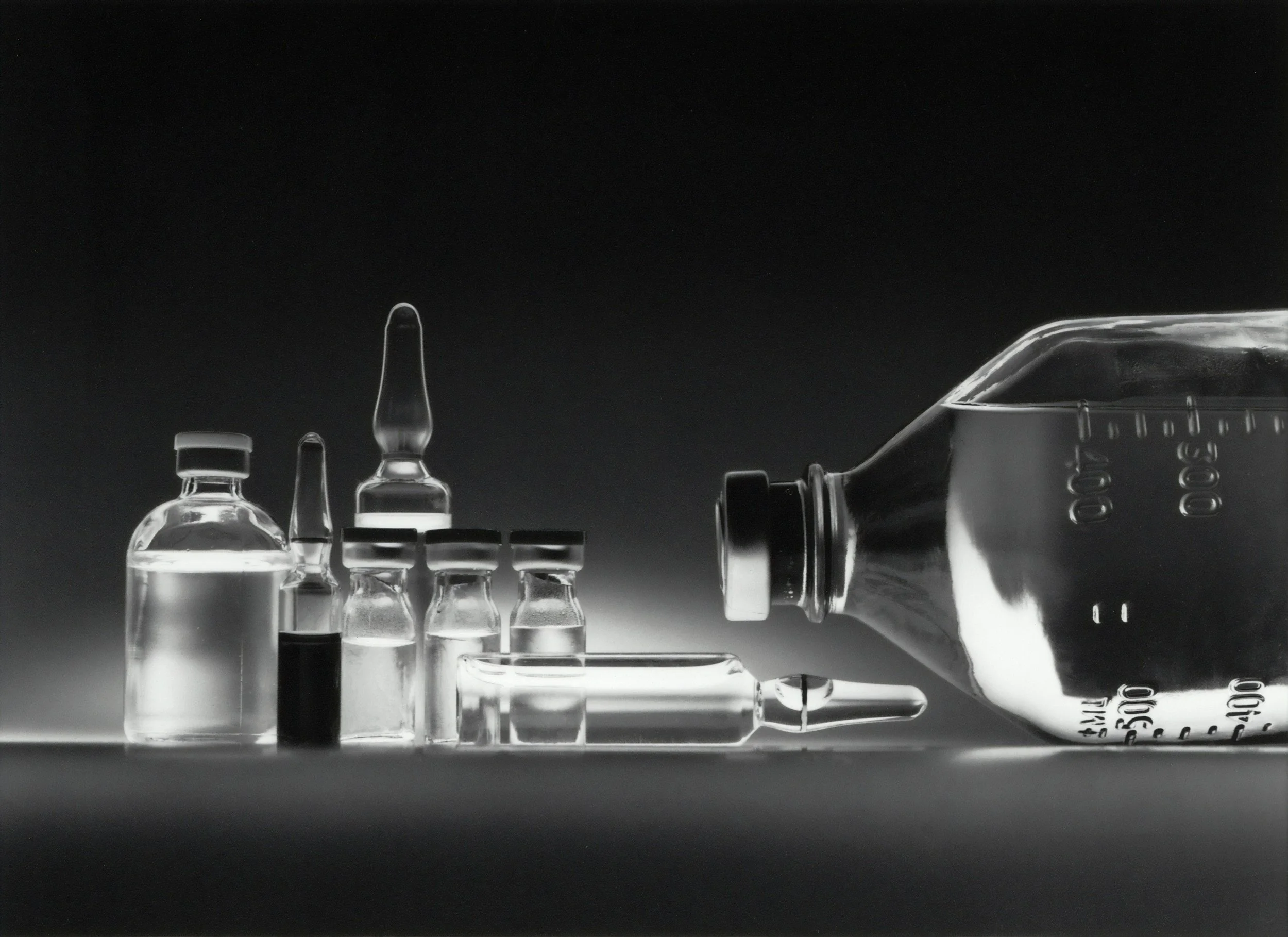 Collection of medical ampoules and vials, with a large glass ampoule lying horizontally and a syringe, all in black and white