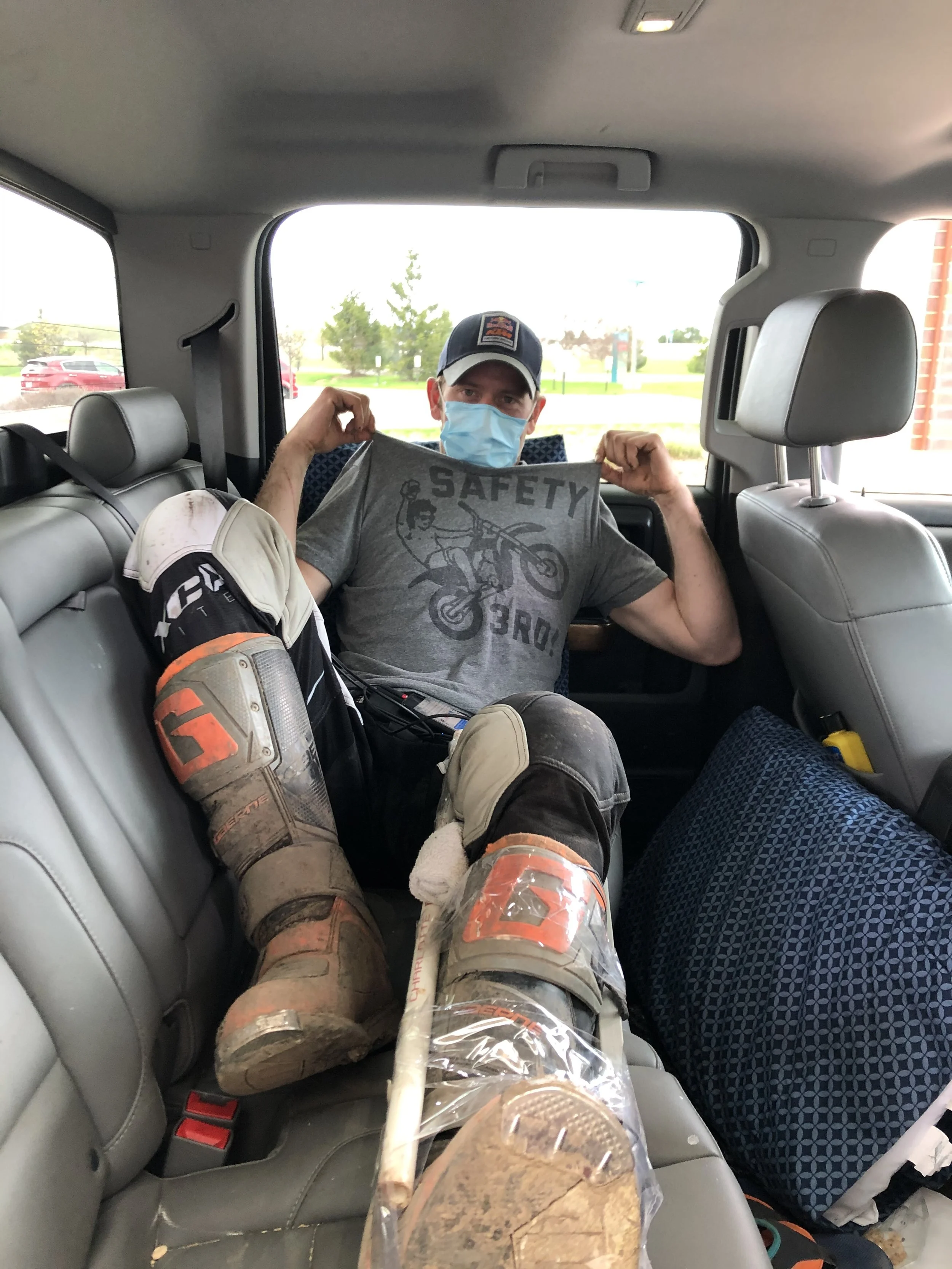 Man wearing a face mask and a baseball cap sitting in the backseat of a vehicle, holding up a gray t-shirt with the word 'SAFETY' and a motocross graphic. He is wearing motocross boots and gear, with a dirt-covered boot resting on the seat.