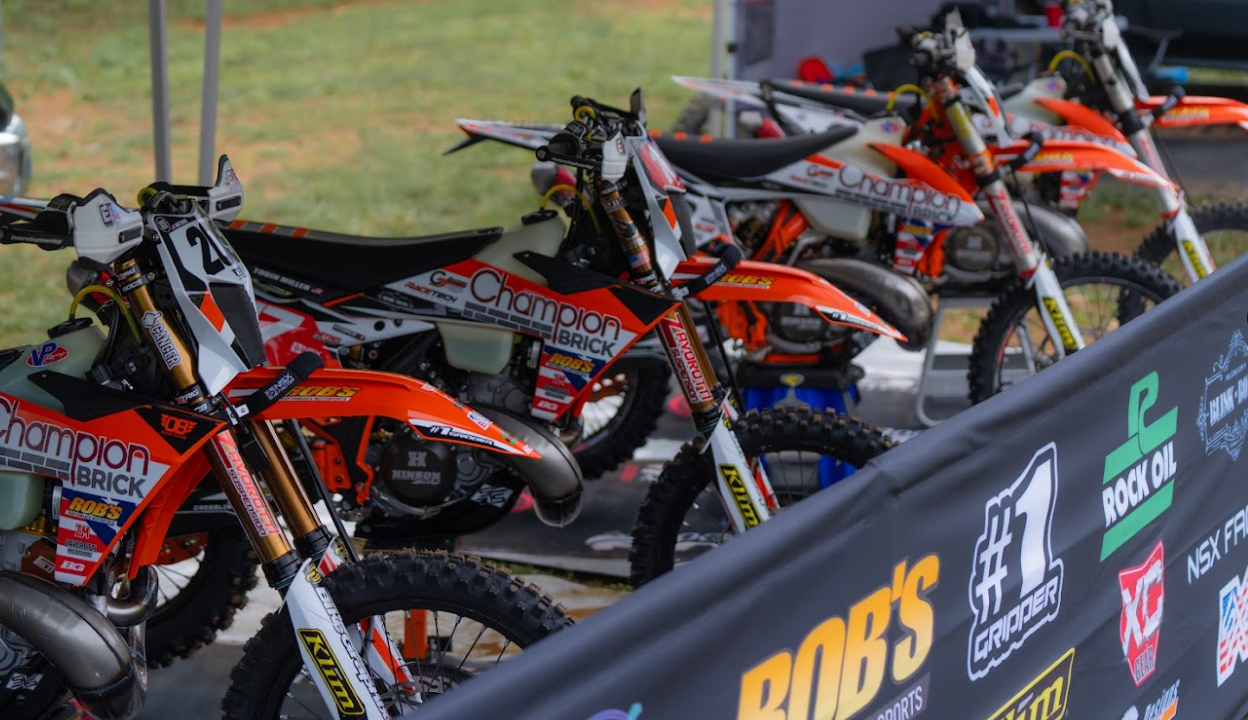 A lineup of orange and white dirt bikes with sponsor decals, parked behind a black barrier with logos at a motocross event.