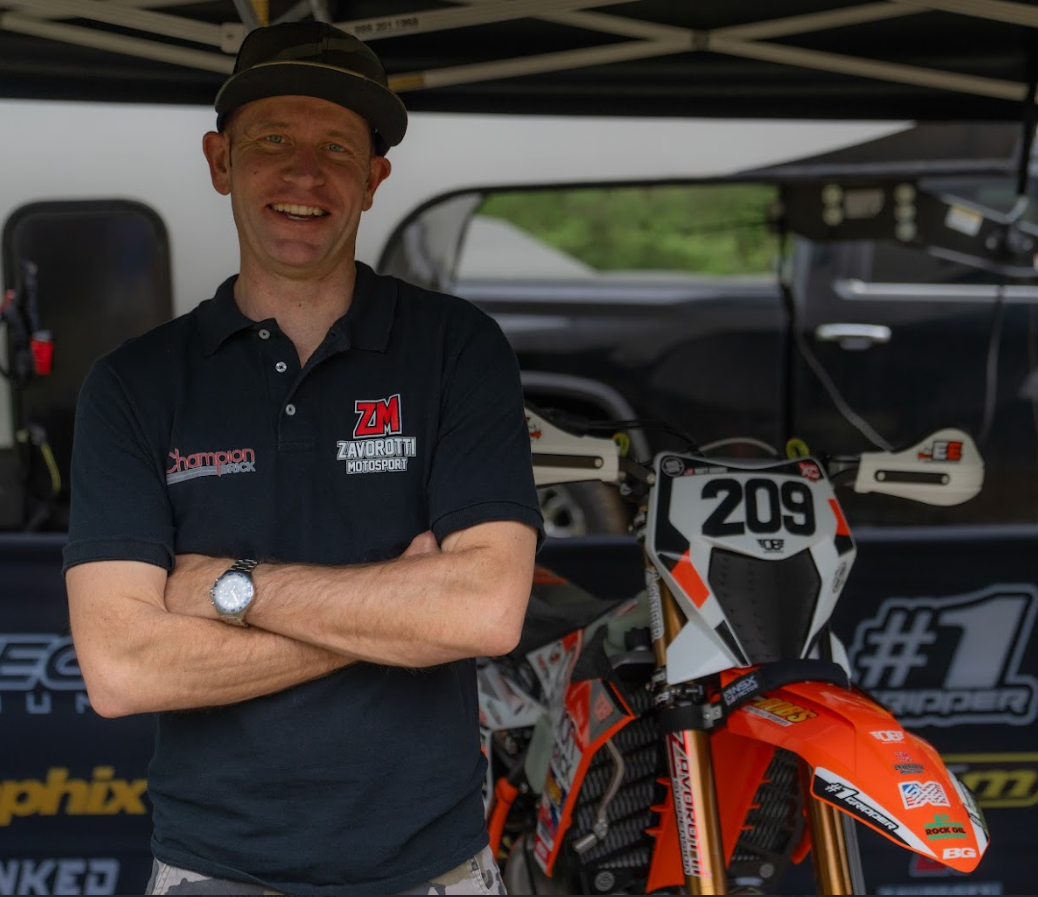 A man standing in front of a racing motorcycle inside a tent. He is wearing a black polo shirt with racing team logos and a watch, smiling with arms crossed.