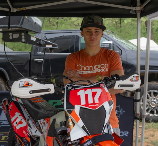 Teenage boy with crossed arms wearing an orange t-shirt and a baseball cap, standing behind a motocross dirt bike with number 117, under a canopy with a parked truck and a vehicle in the background.