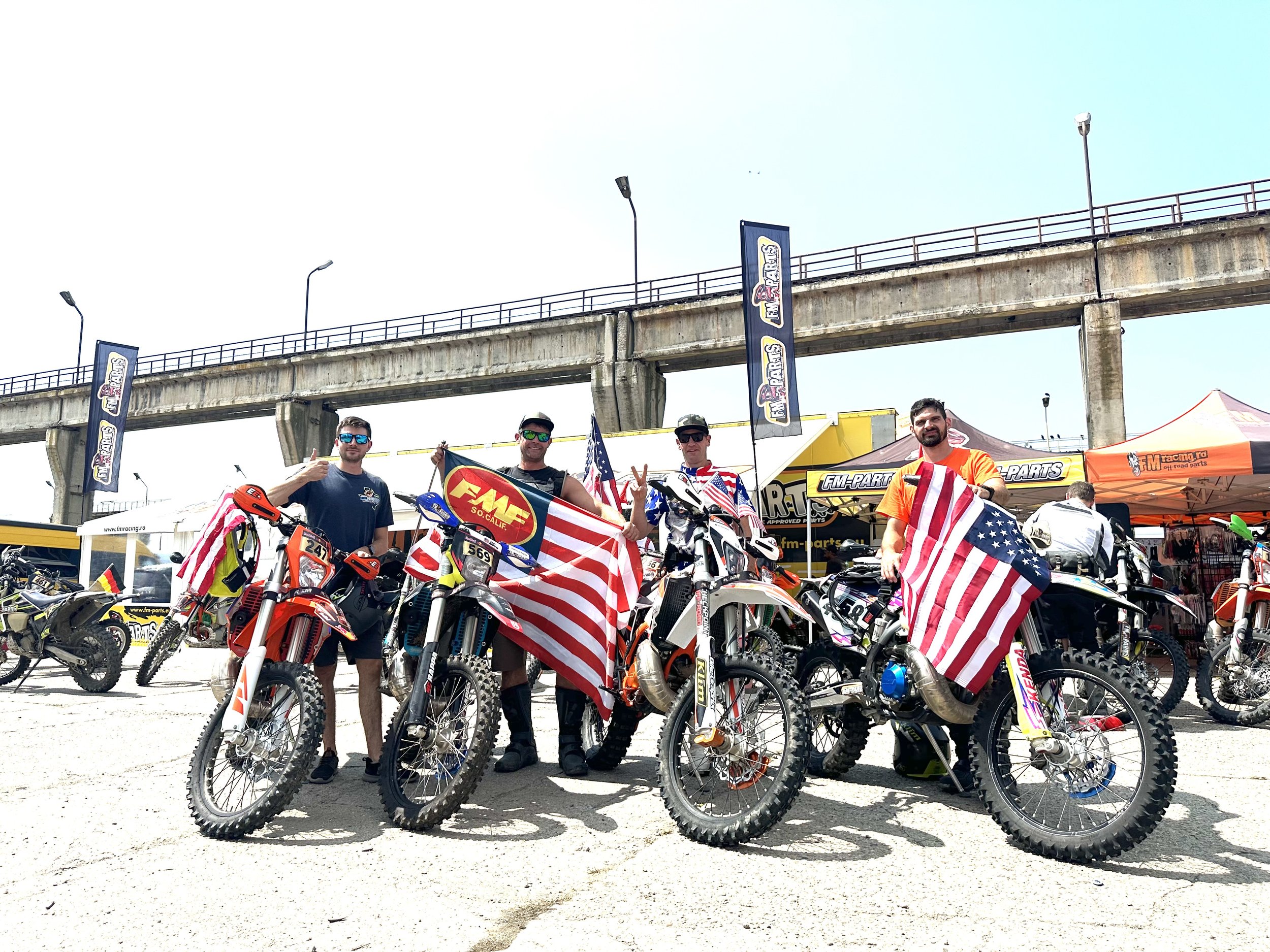 Four men standing next to motocross bikes holding American flags, in a dirt area with tents and banners, under a concrete overpass on a sunny day.