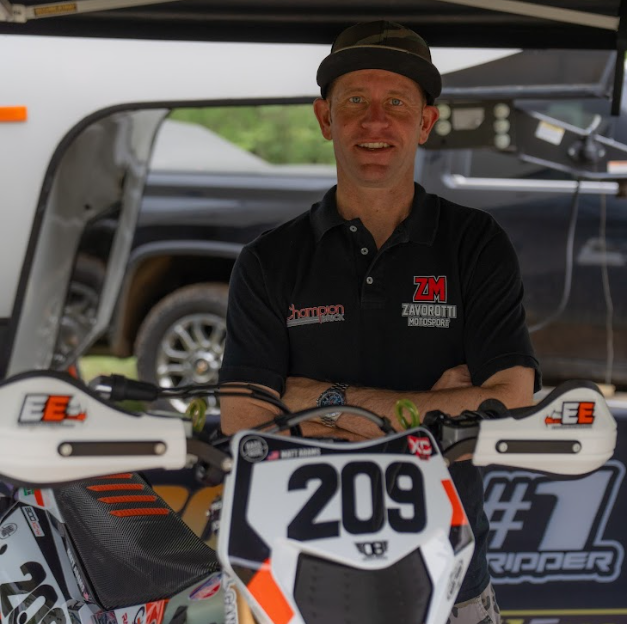 A man smiling and standing behind a dirt bike numbered 209, at an outdoor event, with a vehicle and canopy in the background.