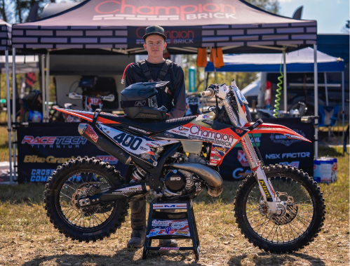 A young motocross rider standing behind his dirt bike at a racing event, with a tent in the background displaying sponsors' logos.