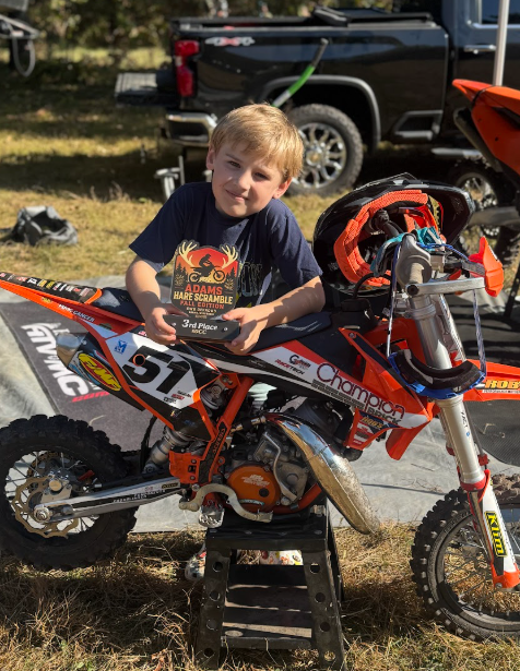 A young boy sitting on a dirt bike holding a trophy. There is a black pickup truck in the background.