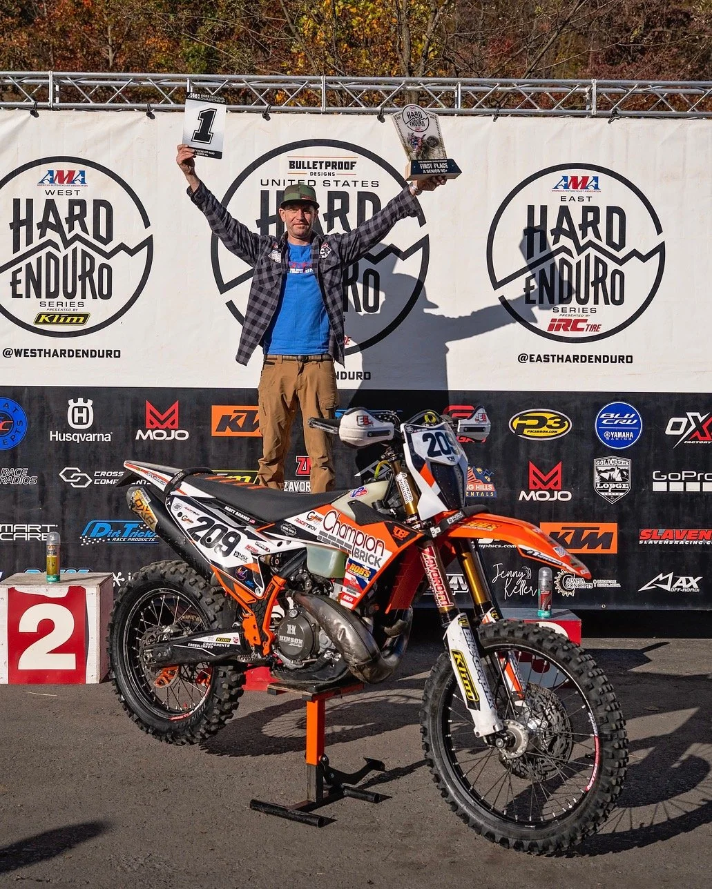 A man standing on a podium with his arms raised, holding a trophy and a sign that says "1st" in front of a backdrop with "Hard Enduro Series" logos. In front of him is a dirt bike with the number 209, and the scene is set outdoors with autumn trees in the background.
