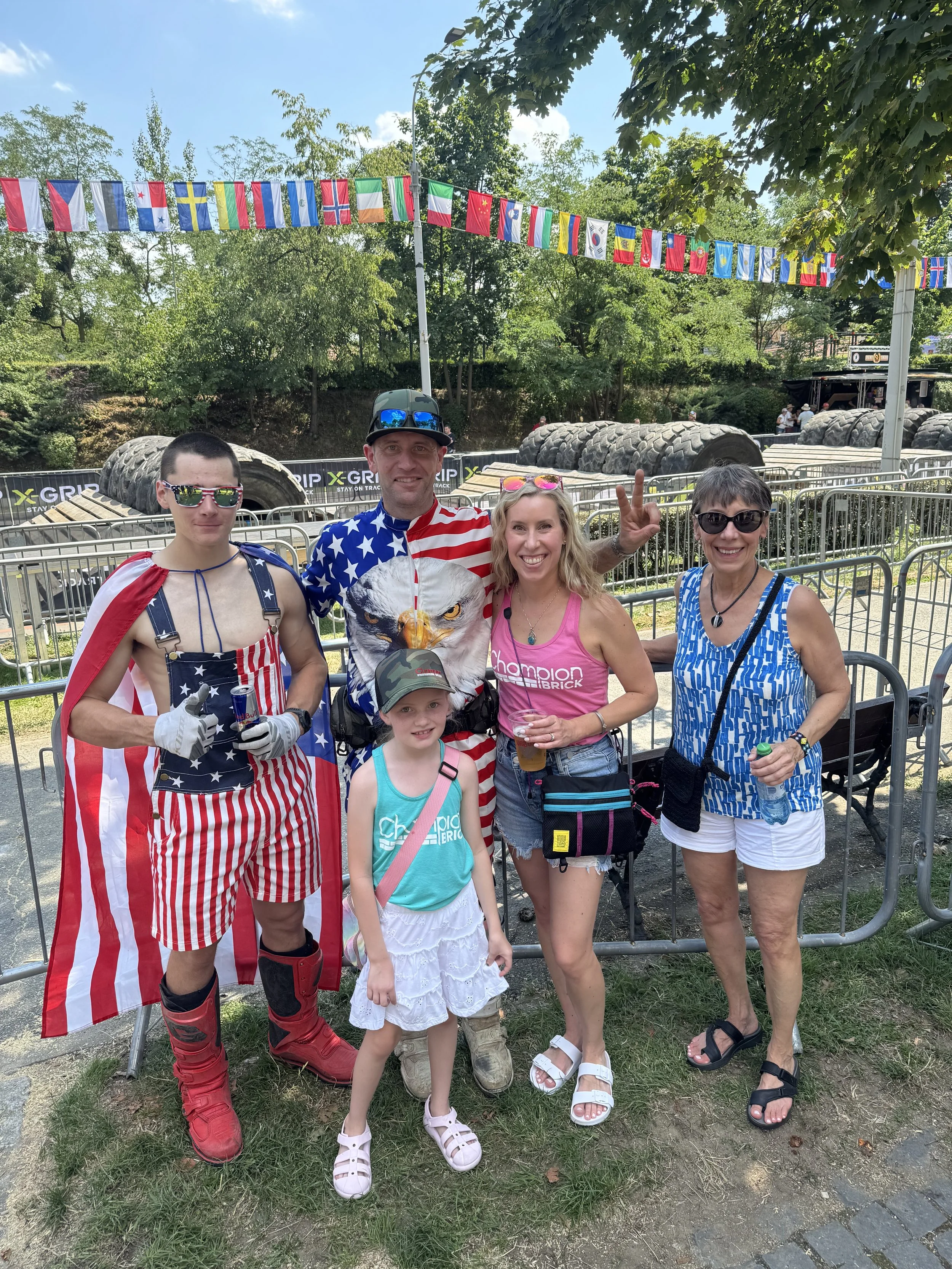 Group of five people dressed in patriotic American clothing, standing outdoors at an event with flags and tires in the background.