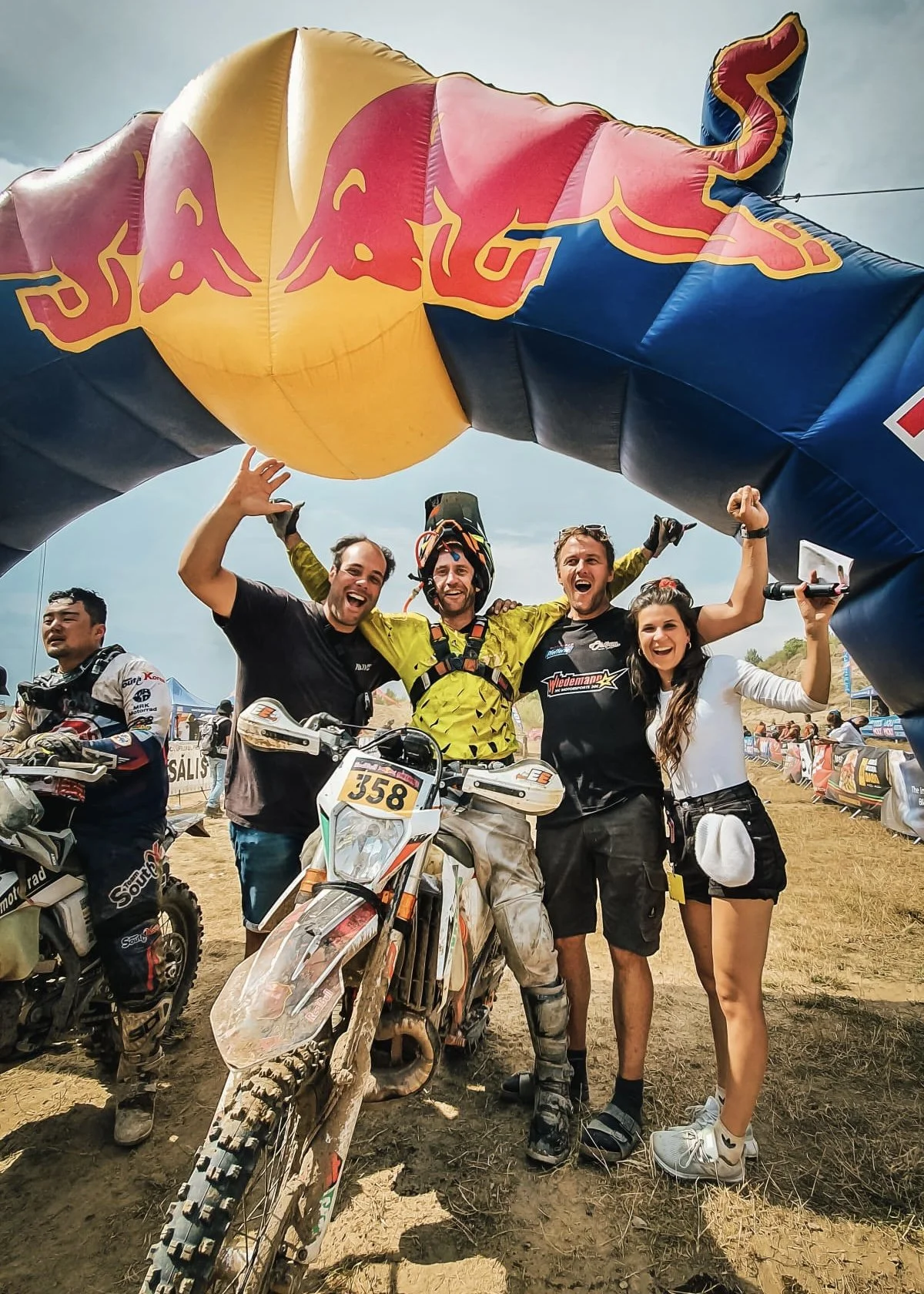 A group of five people celebrating under a Red Bull arch at a motocross event, with dirt bikes and a dirt track in the background.