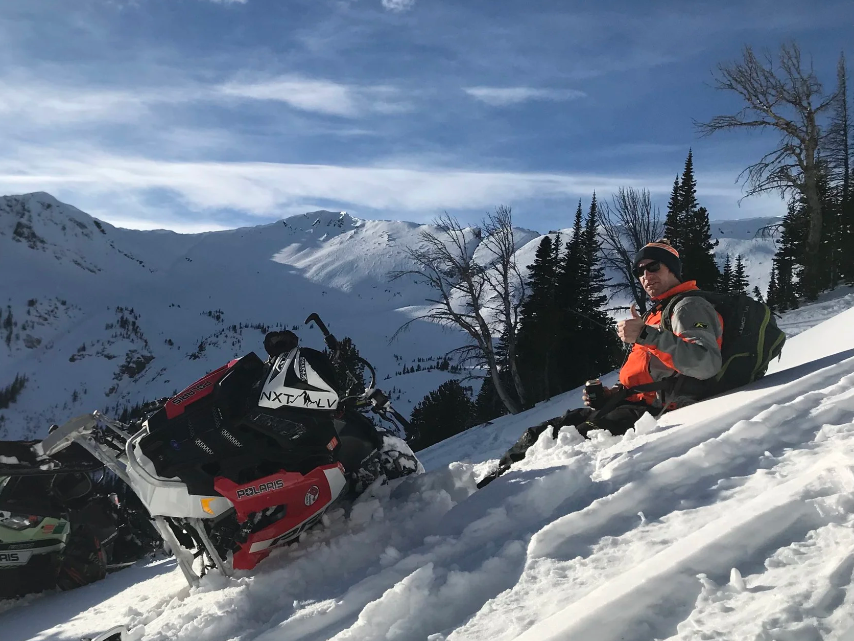 A man in snow gear sitting on snowy mountainside with a backpack, holding a mug, next to a parked snowmobile in a snowy mountain landscape with pine trees and a blue sky.