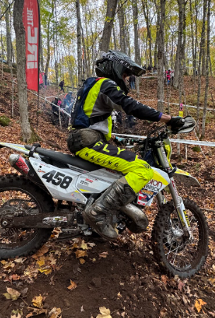 Motorcyclist wearing a helmet and yellow riding gear navigating a dirt trail in a wooded area, with spectators and a red banner in the background.