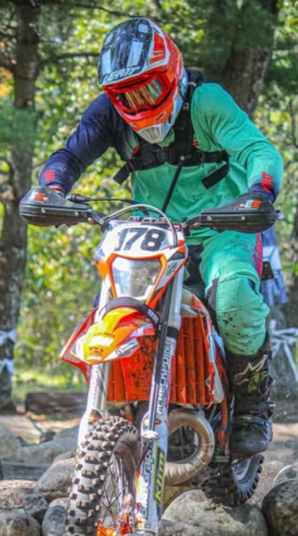 Motocross rider wearing a red helmet, teal and navy gear, riding an orange and white dirt bike over rocks in a forest trail.