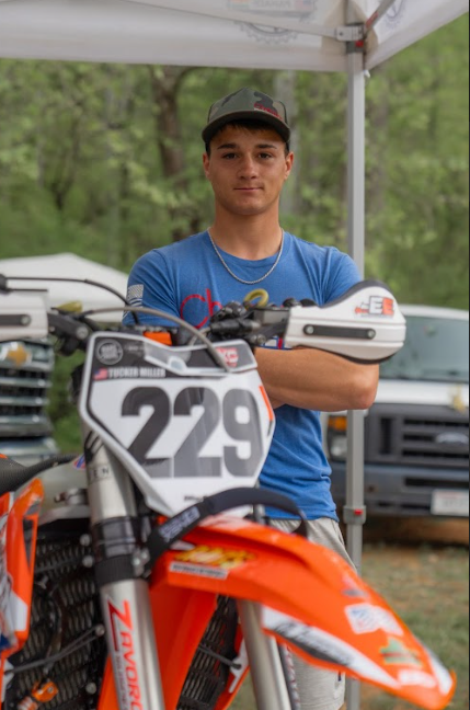 Young man standing behind an orange dirt bike with the number 229, wearing a blue t-shirt and a black cap, outdoors under a canopy with trees in the background.