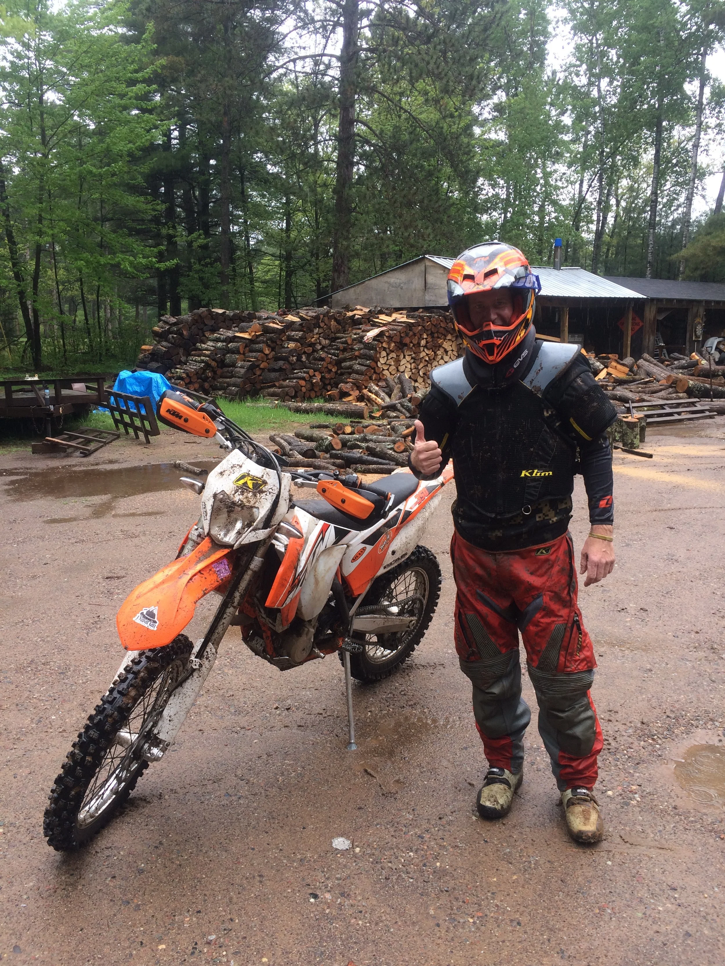 A person in motocross gear, including a helmet, goggles, and protective clothing, standing next to an orange dirt bike on a muddy outdoor trail, giving a thumbs-up.