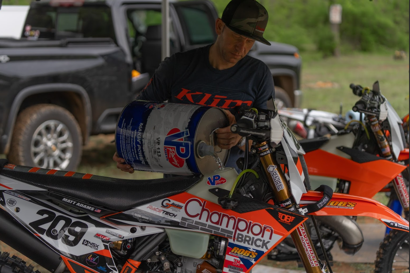 A man filling a dirt bike with VP racing fuel from a large canister. The dirt bike is orange and white with various sponsor stickers, and he is wearing a dark KTM shirt and a cap. There is another similar dirt bike beside him, and a black vehicle parked in the background on the grassy terrain.