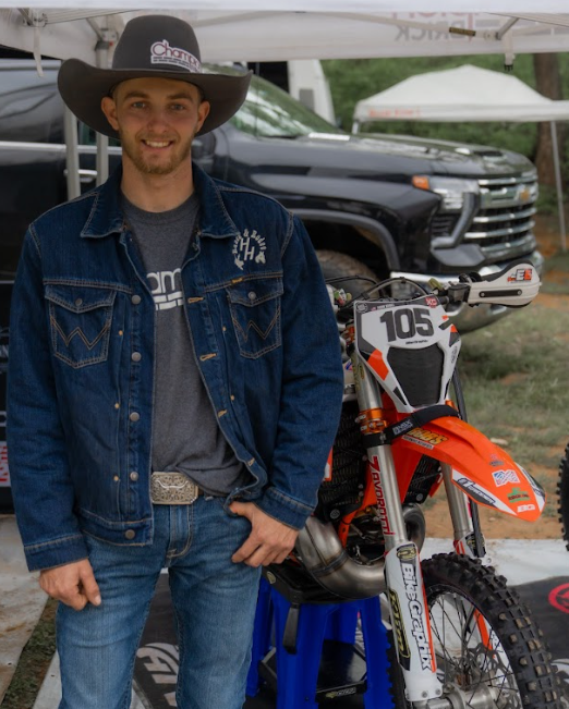 Young man with a beard smiling, wearing a cowboy hat with a logo, denim jacket, and jeans, standing next to a dirt bike with the number 105, under a canopy at an outdoor event.