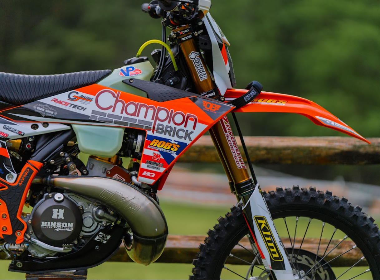 Close-up of a dirt bike with orange, black, and white graphics, featuring sponsor logos, suspension system, and knobby tire, set outdoors with a blurred background of greenery.